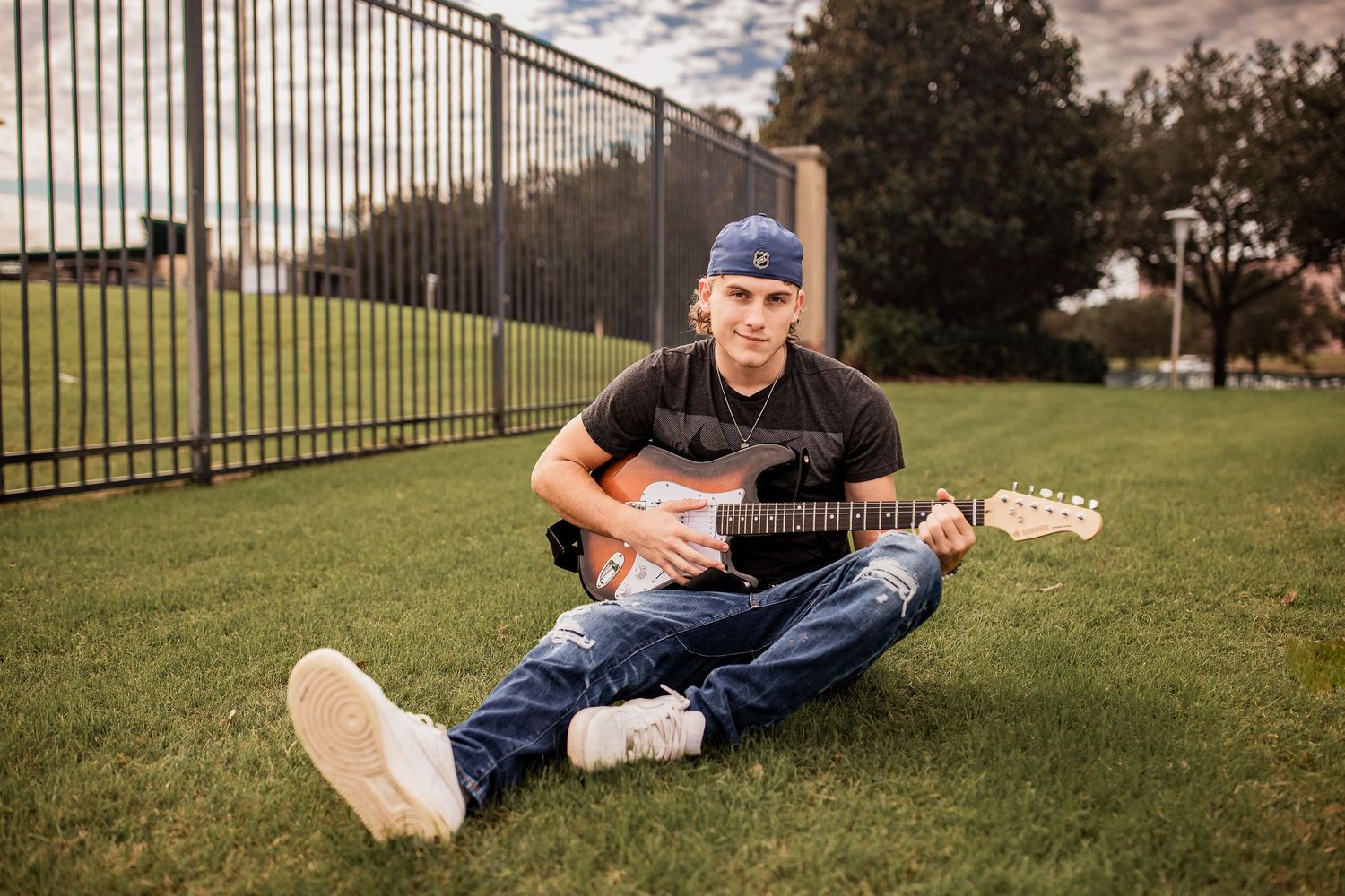 Young man in blue cap sits on green grass playing electric guitar near iron fence at sunset.