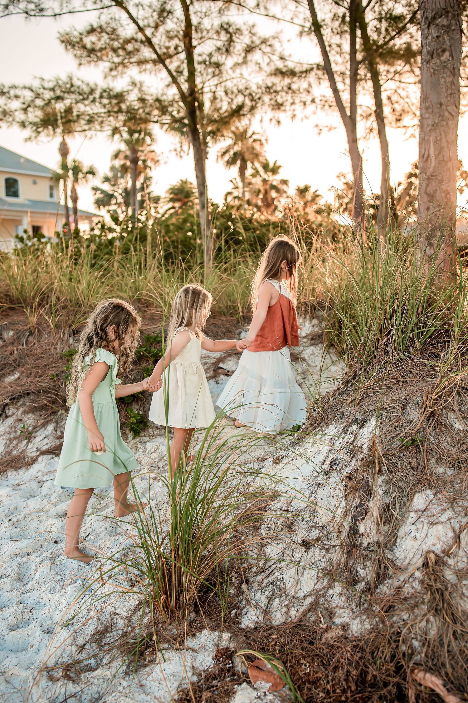 Three girls in pastel dresses walk hand-in-hand through sandy beach dunes at golden hour sunset.