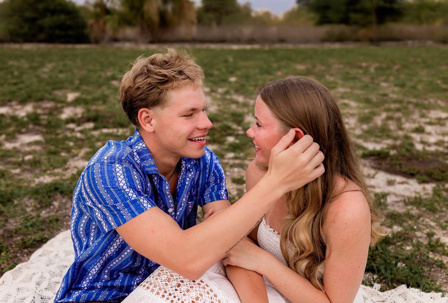 Young couple sitting on blanket in field, smiling and gazing at each other.