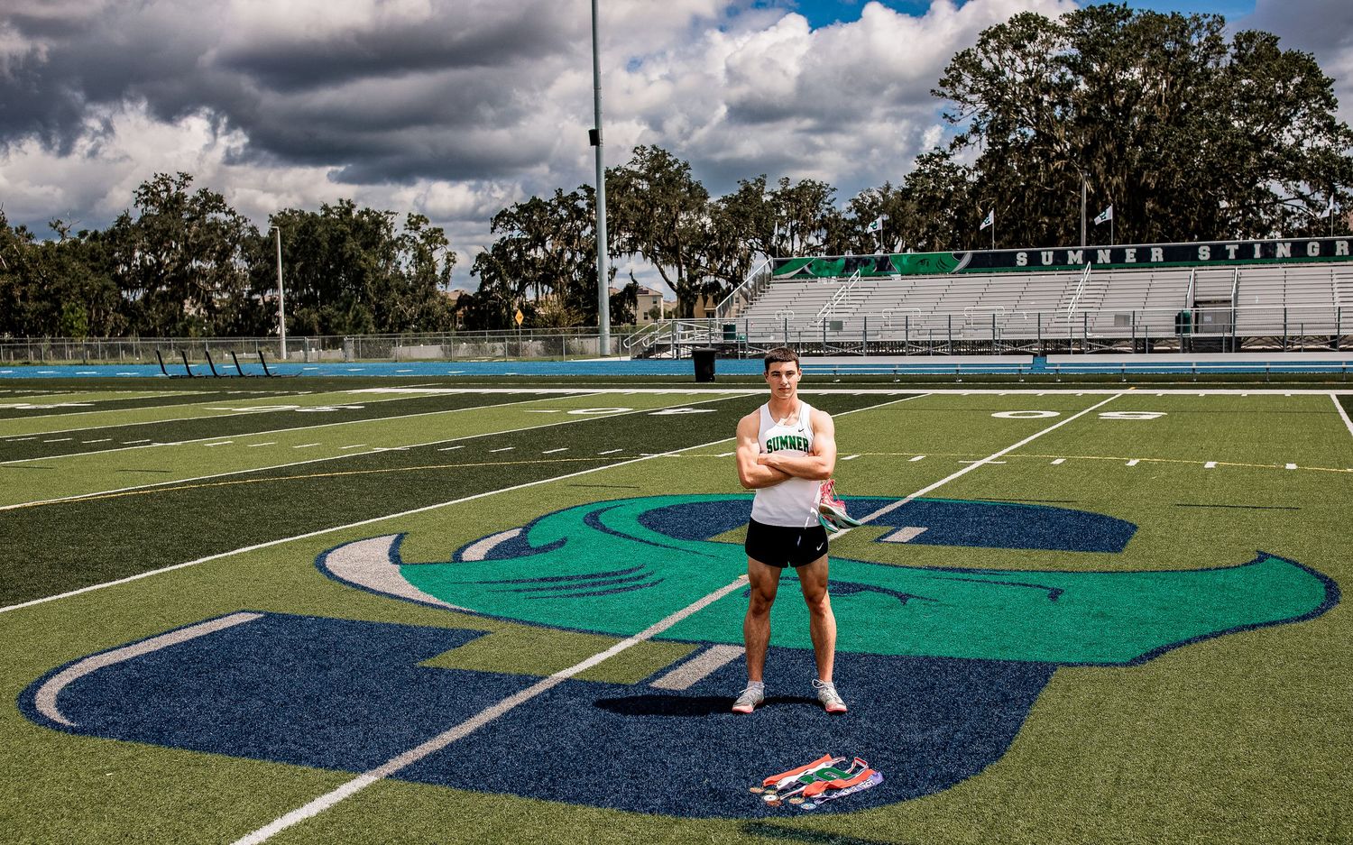 Male athlete stands confidently on a blue and green turf football field with arms crossed during a cloudy day.
