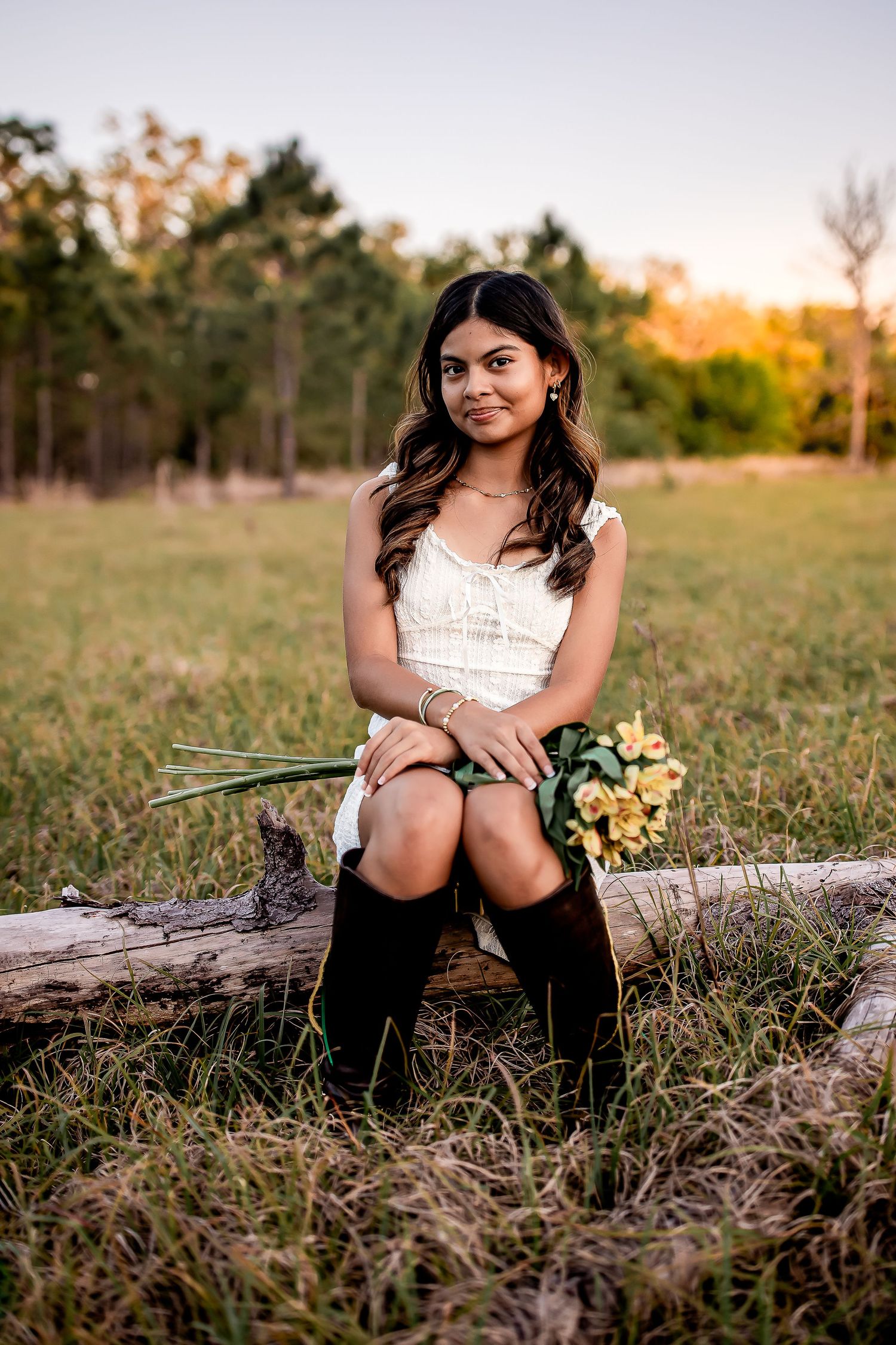 Young woman in white dress and black boots sits on log holding yellow flowers in golden meadow.