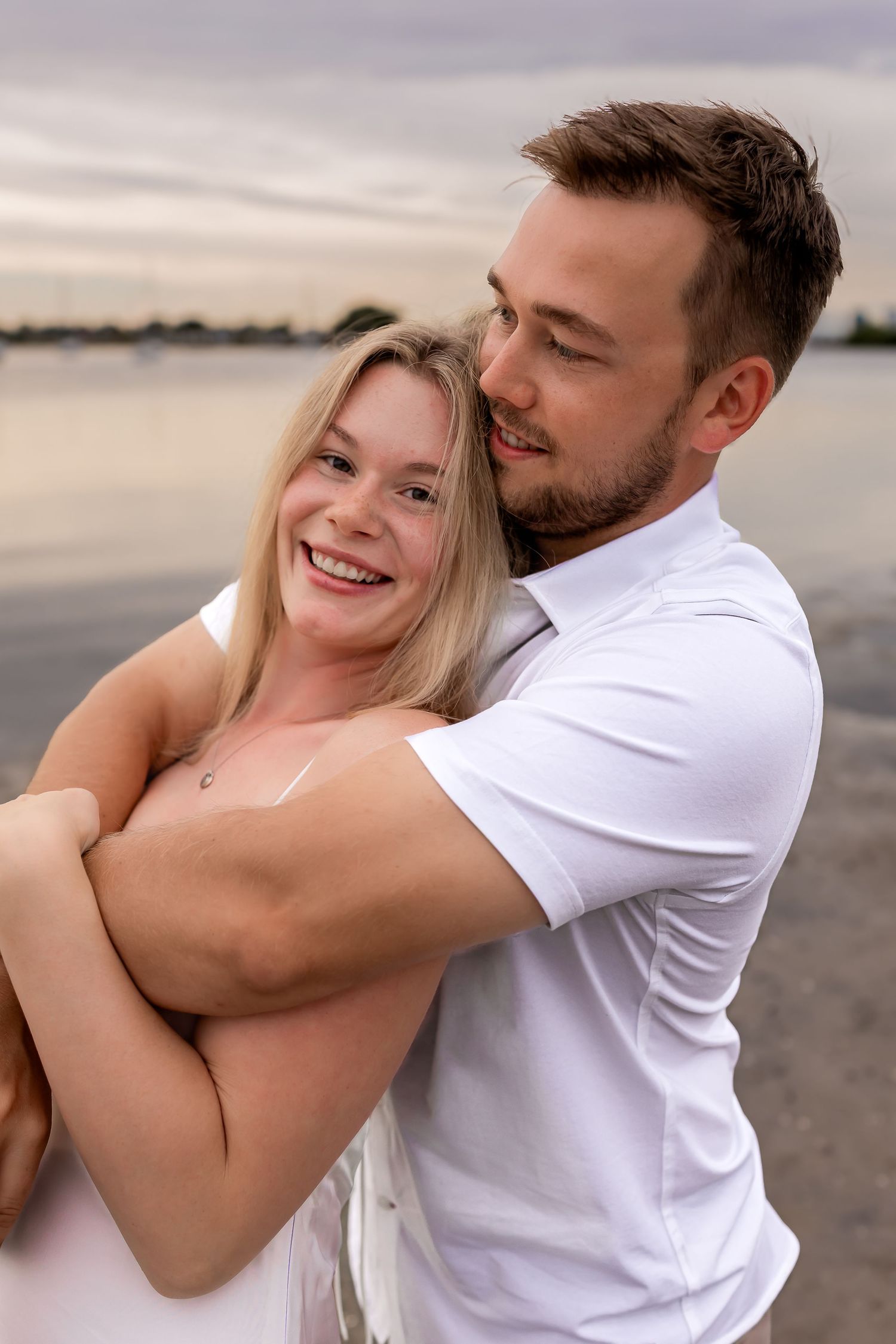 Happy blonde woman smiles as man in white shirt embraces her from behind at waterfront.