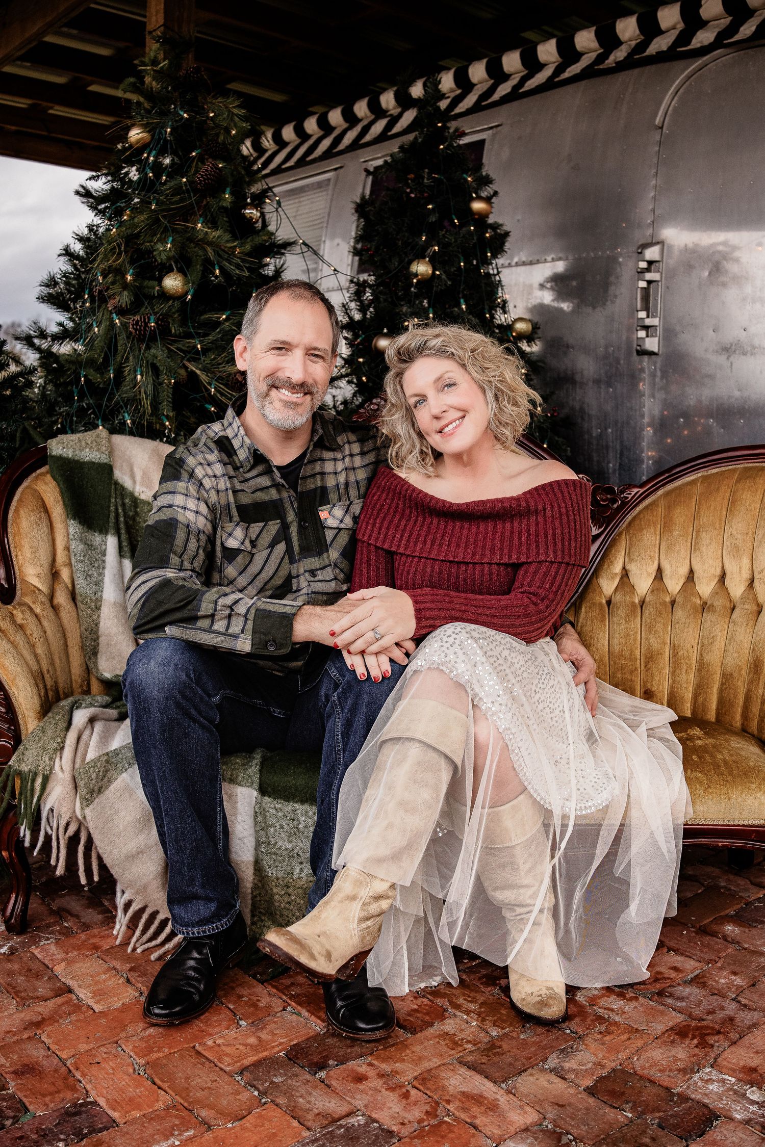 Couple sitting together on a vintage sofa with Christmas trees in a festive outdoor setting.