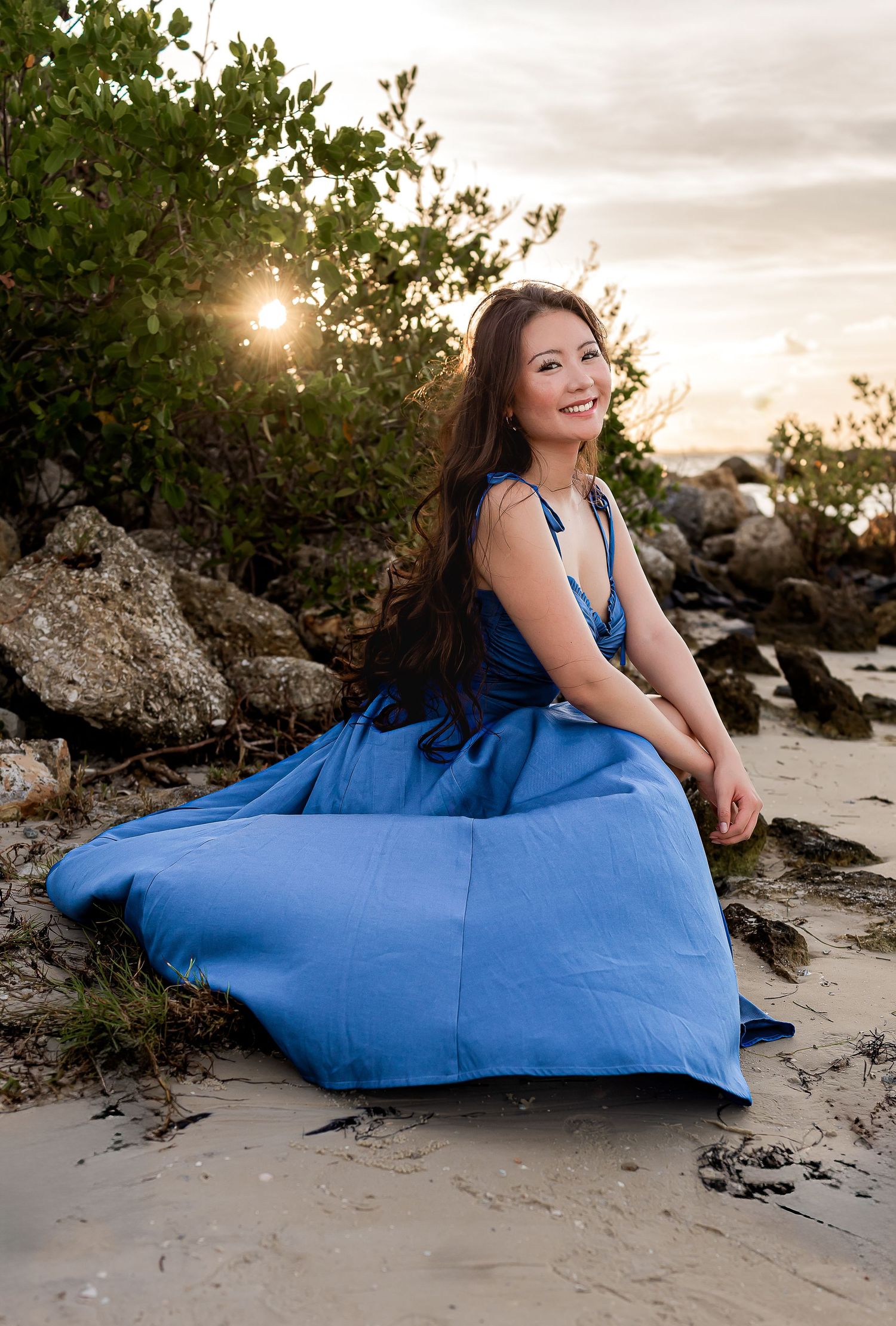 A young woman in a blue gown sits on a sandy beach at golden hour sunset.