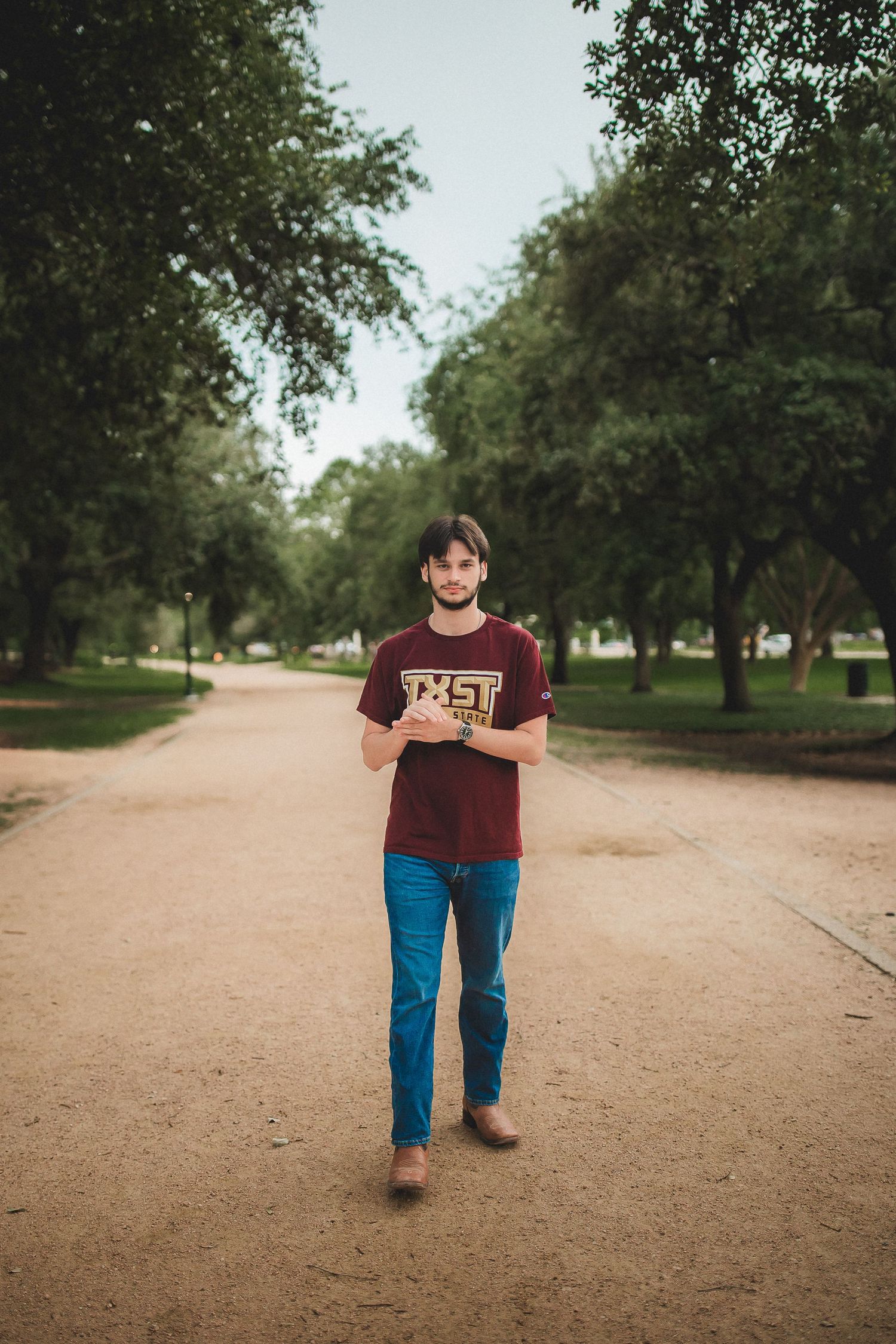 Young man in a maroon TXST shirt walking along a tree-lined park pathway.