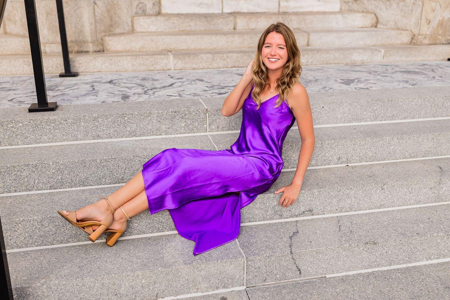 Young woman in purple satin gown sitting on stone steps smiling.