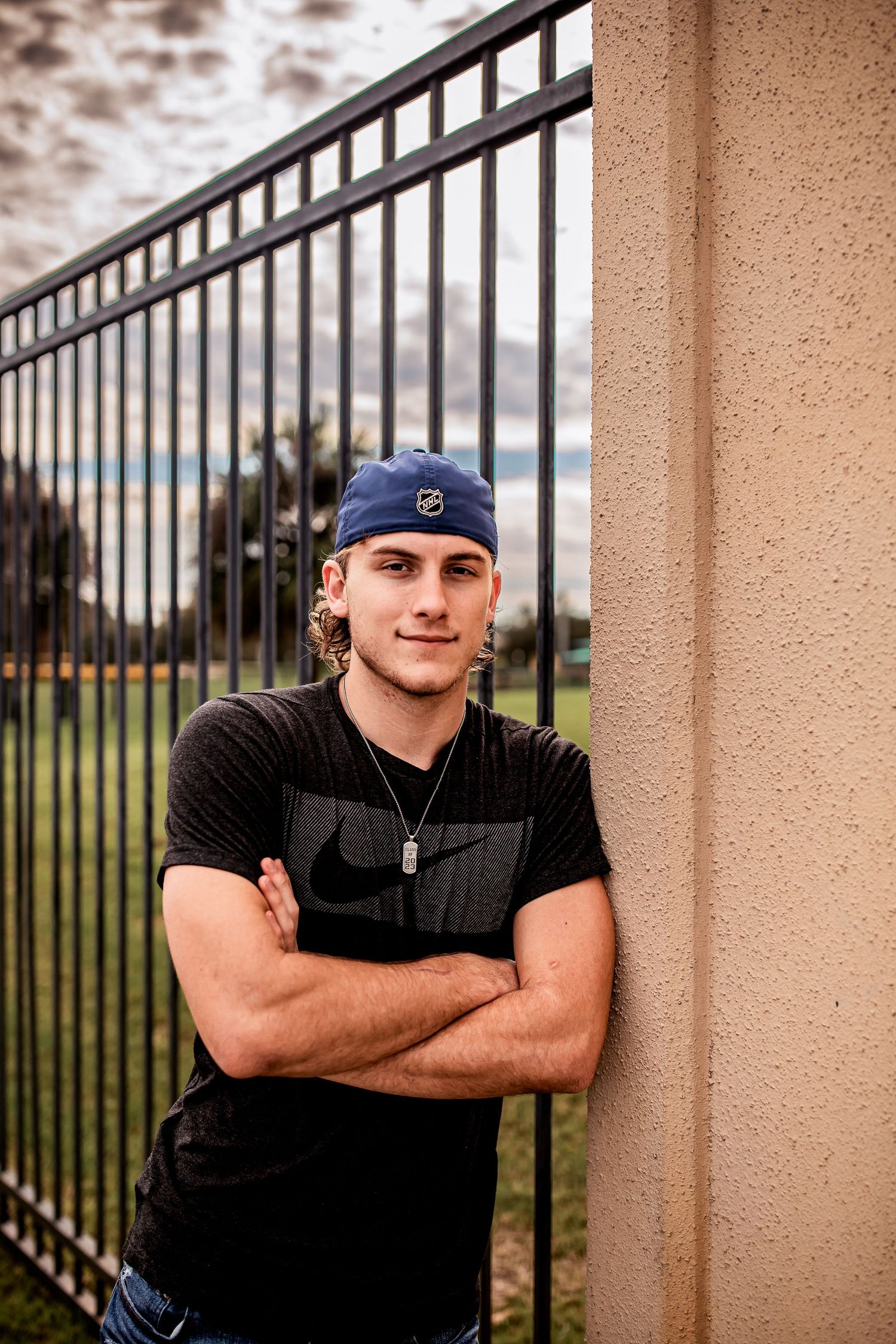Young man in black Nike shirt and blue cap leans against wall with arms crossed near iron fence.