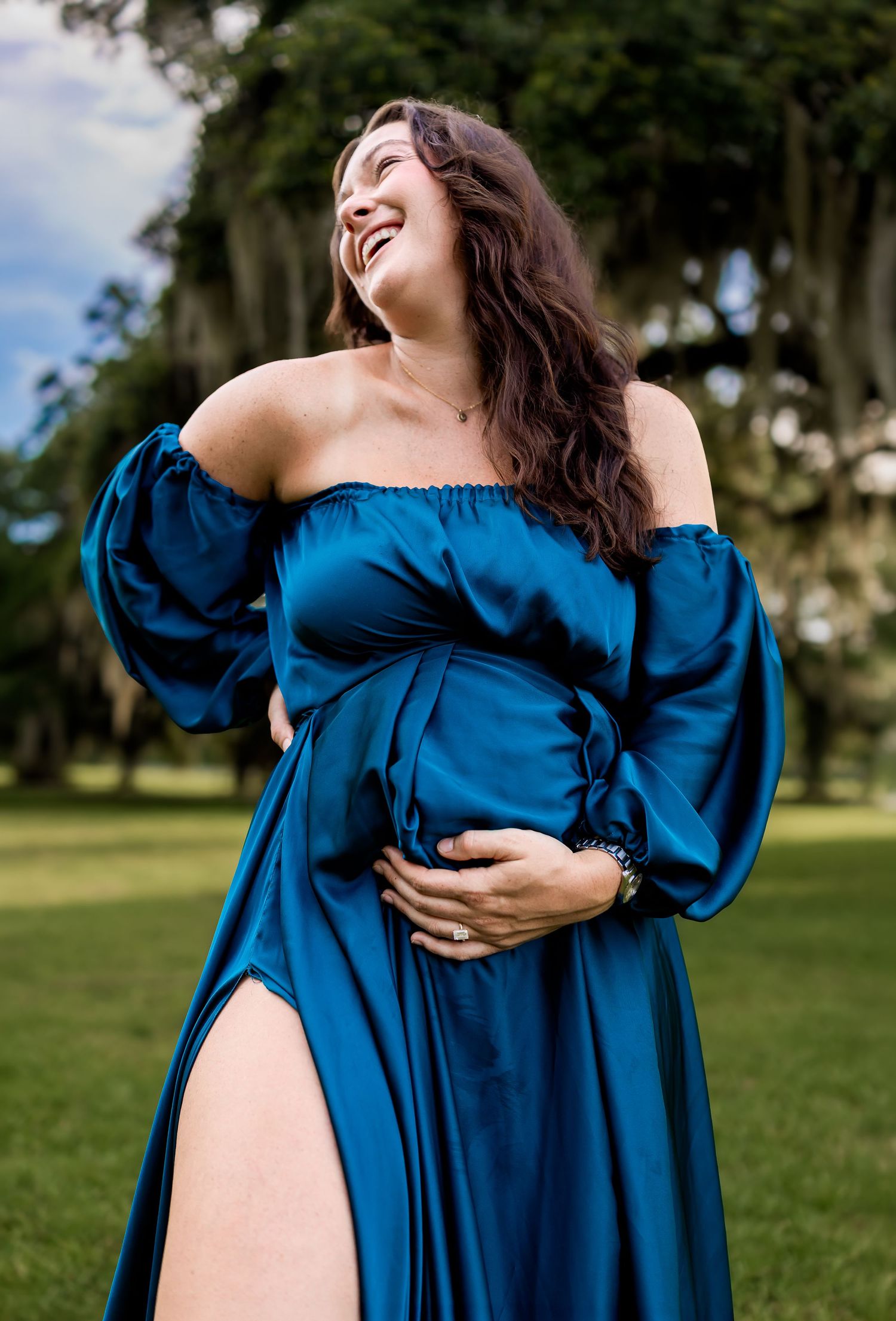 A radiant pregnant woman in a teal off-shoulder satin dress laughing joyfully in a green park.