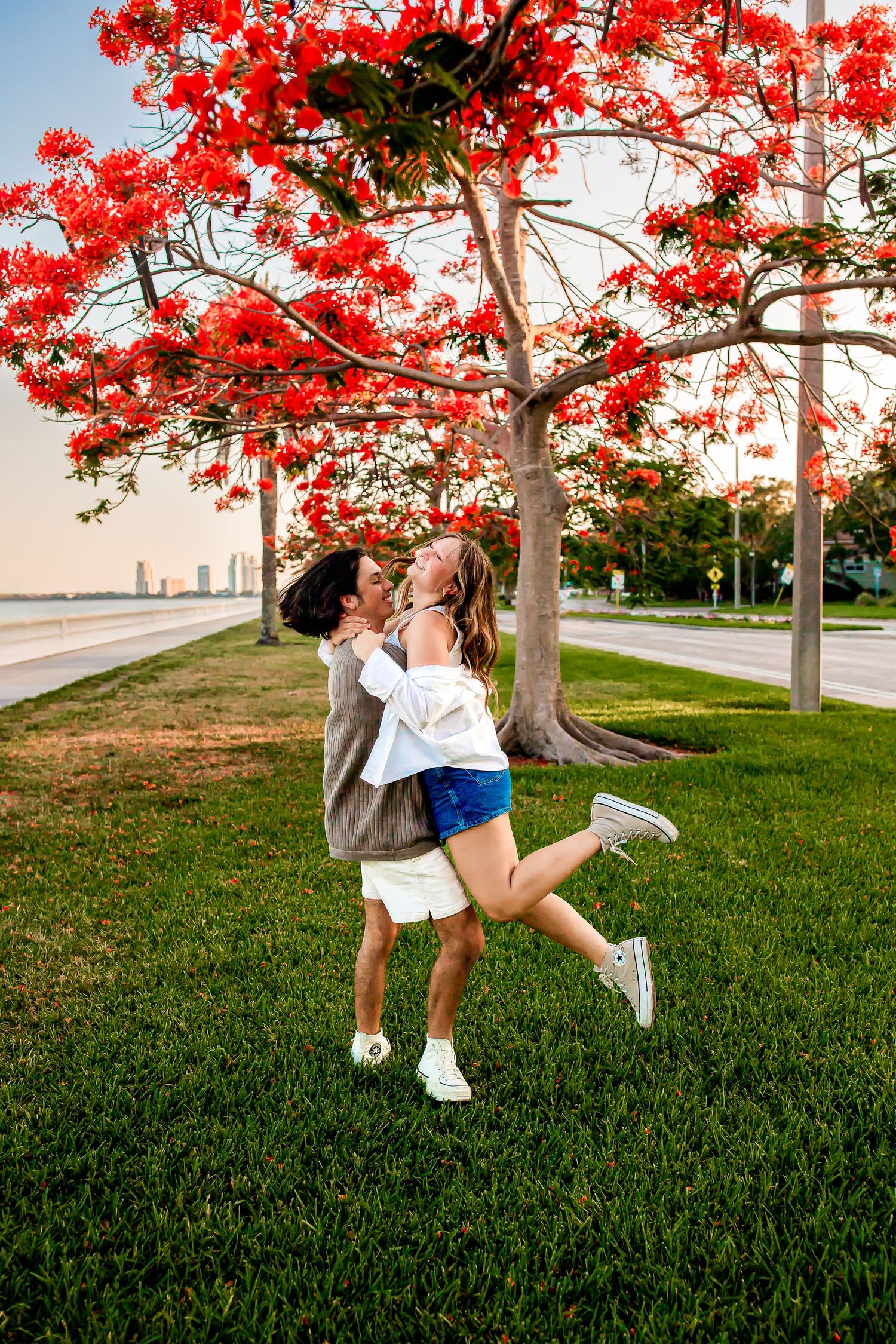 Couple joyfully embraces beneath a stunning red flowering tree on a green lawn at golden hour.