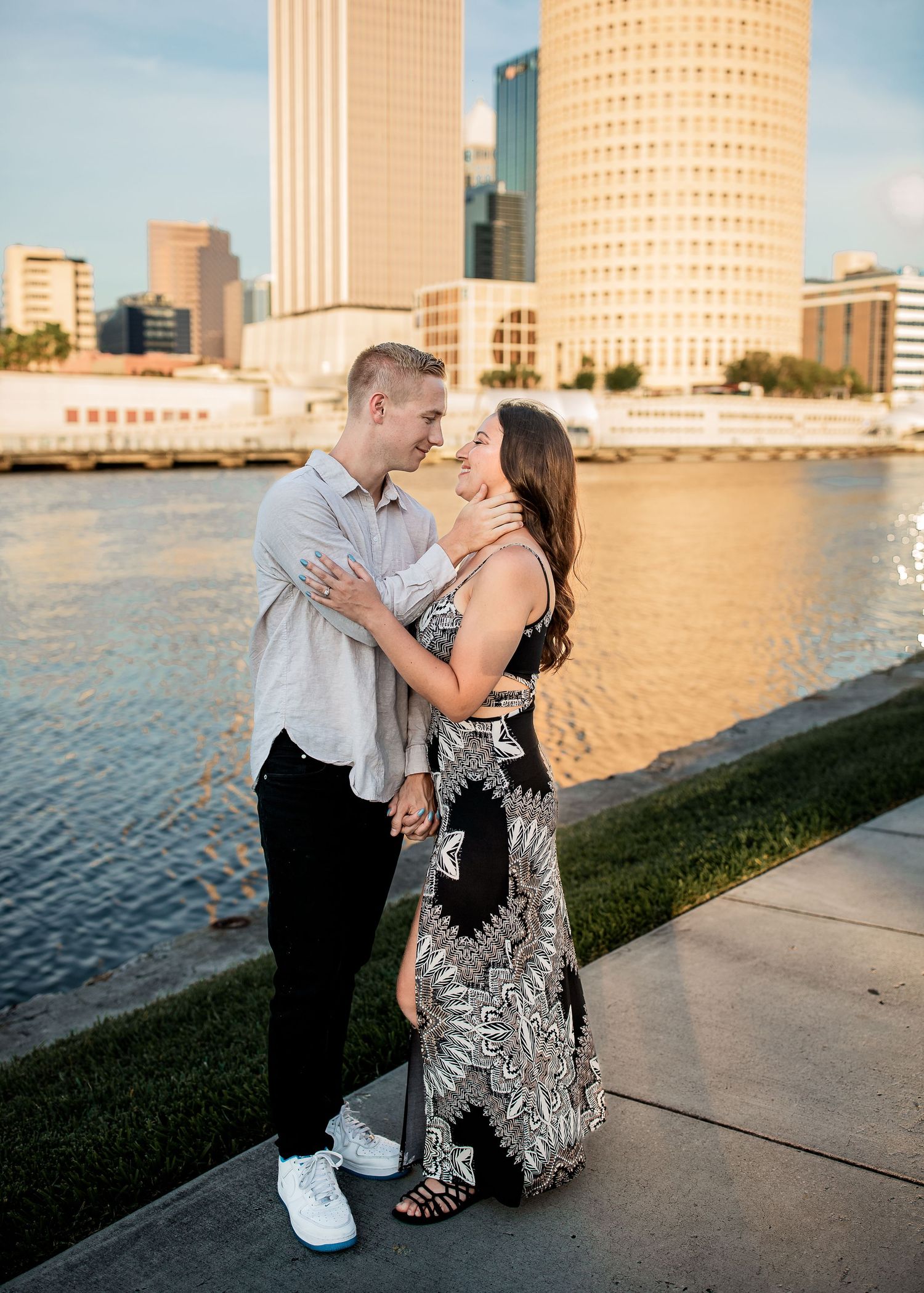 Couple embracing near a waterfront with a stunning city skyline backdrop at sunset.