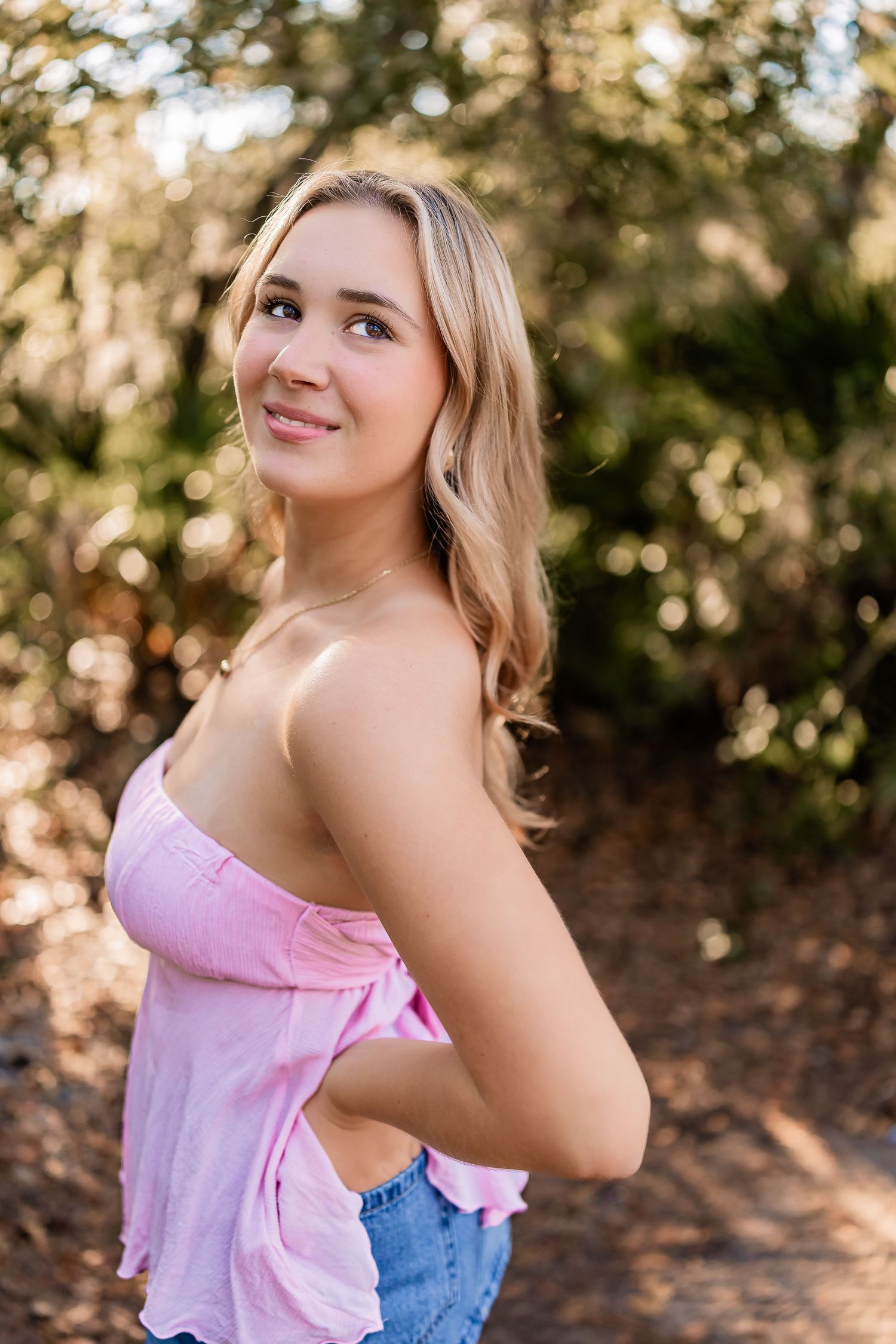 Young blonde womanl in pink off-shoulder top poses outdoors in sun-dappled wooded setting.