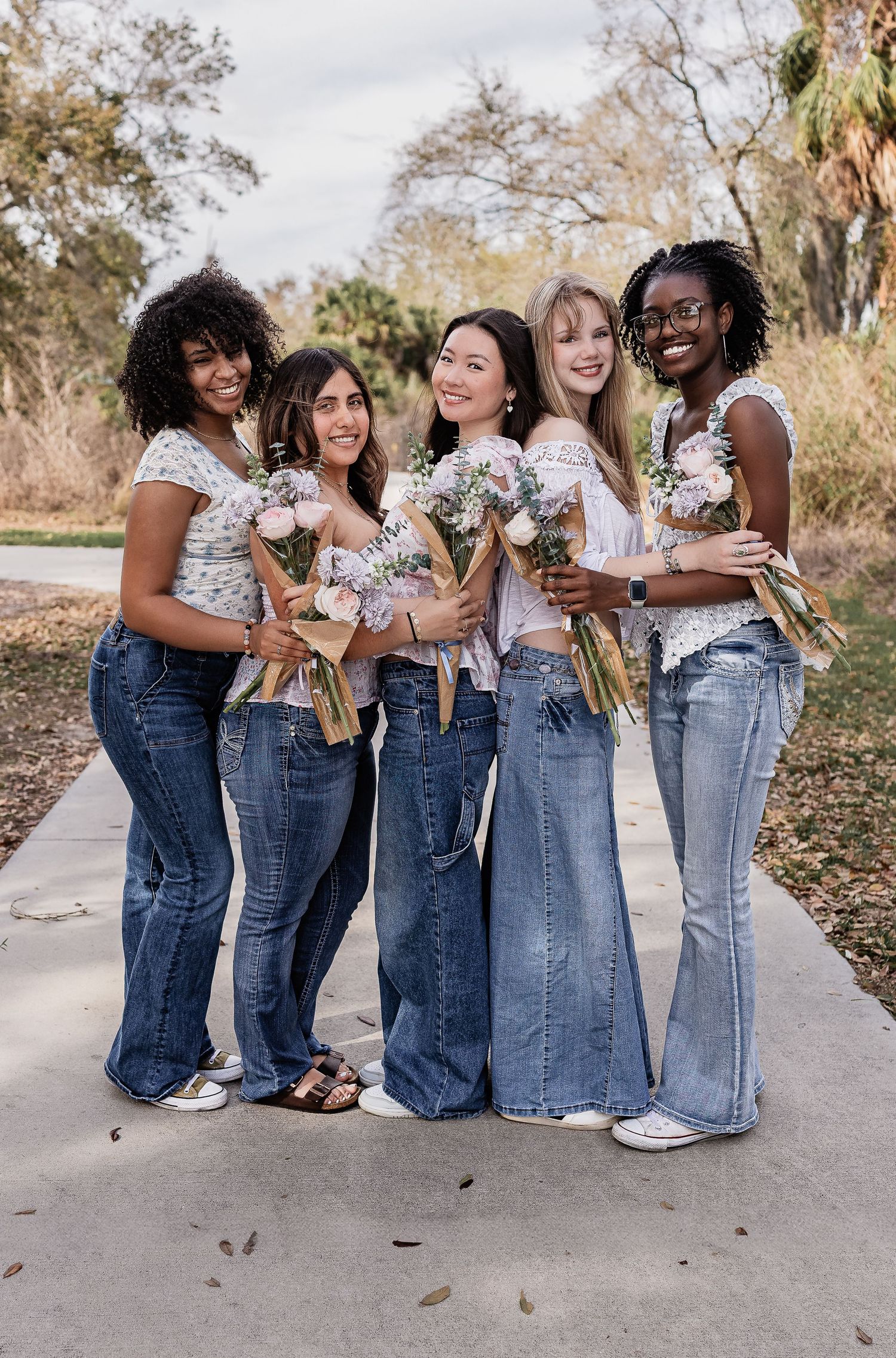 Five diverse young women in white tops and flare jeans holding flower bouquets on an outdoor path.