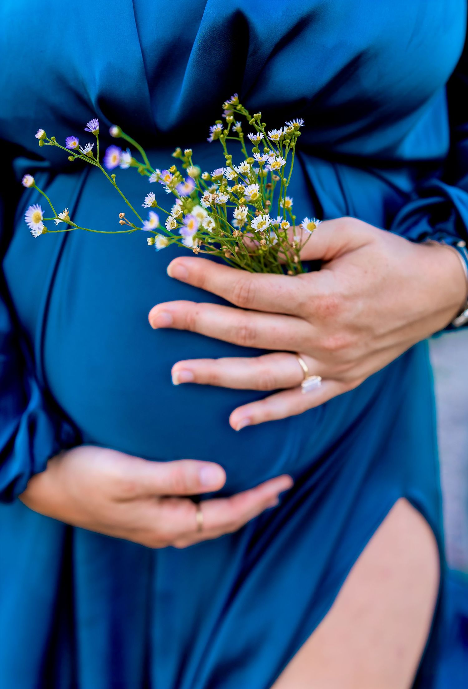 Close-up of pregnant woman in blue dress cradling her belly with wildflowers and a diamond ring.