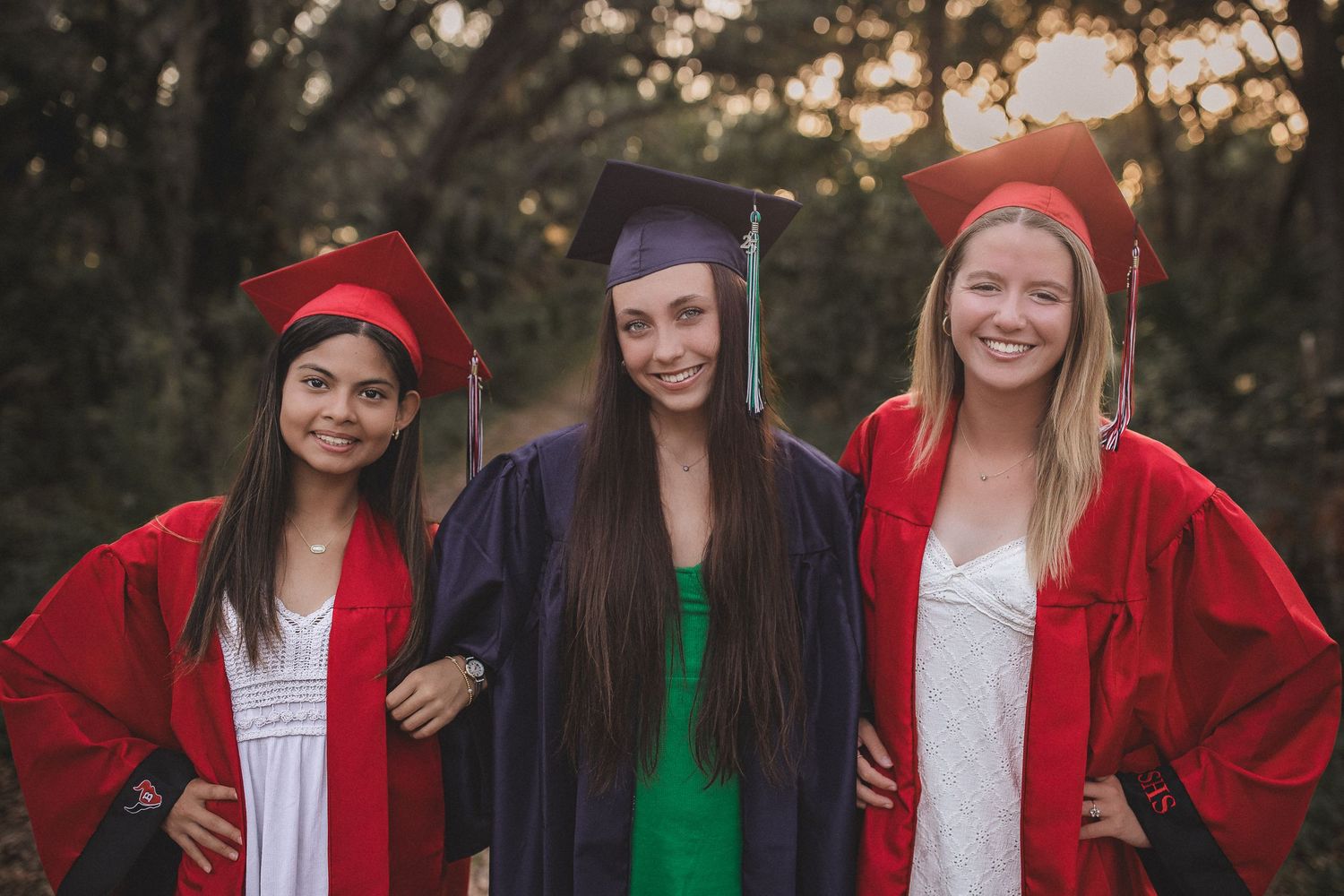 Three high school graduates in red and black caps and gowns posing together outdoors at sunset.