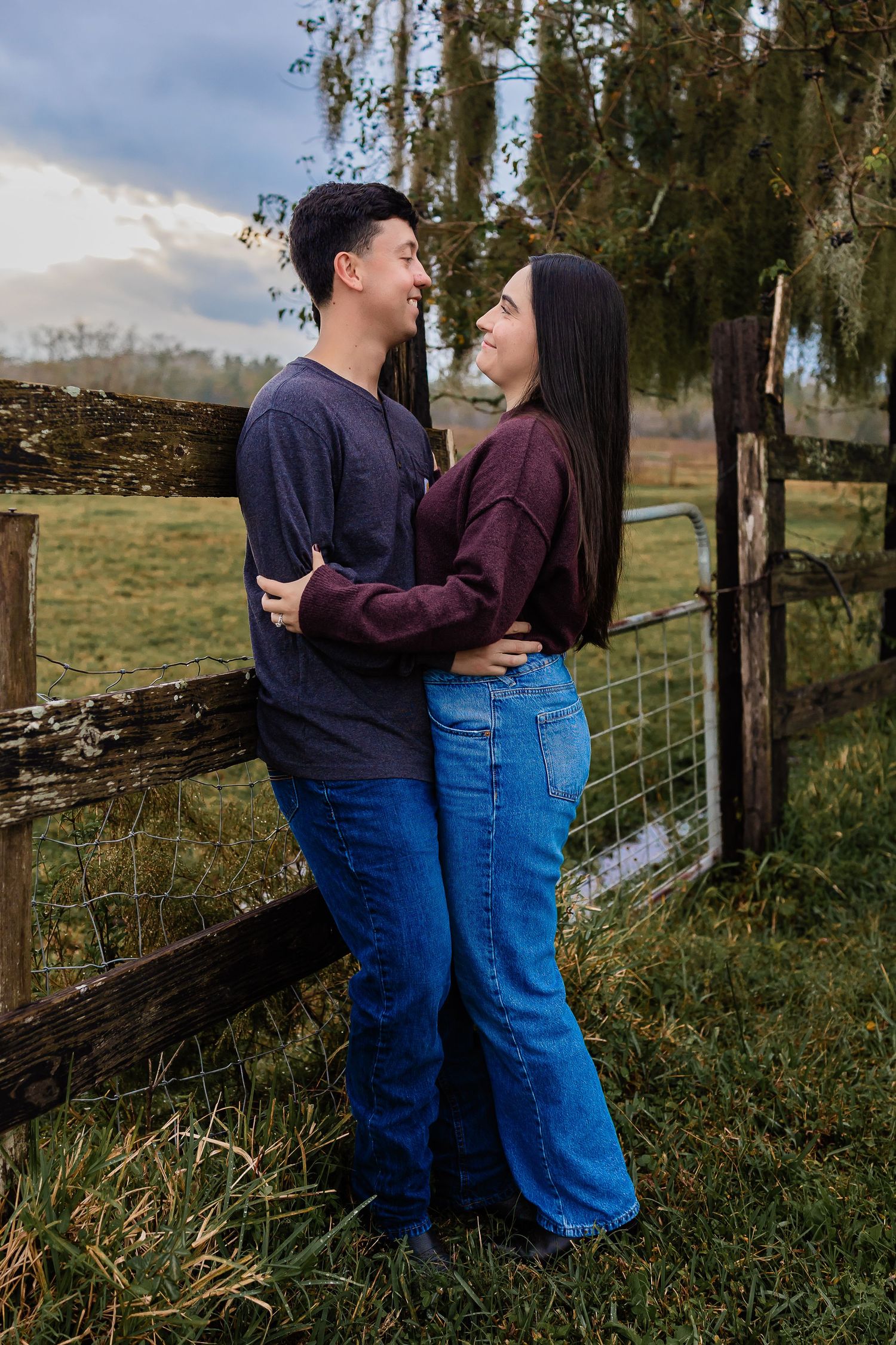 Young couple embracing and smiling at each other by a rustic wooden fence.
