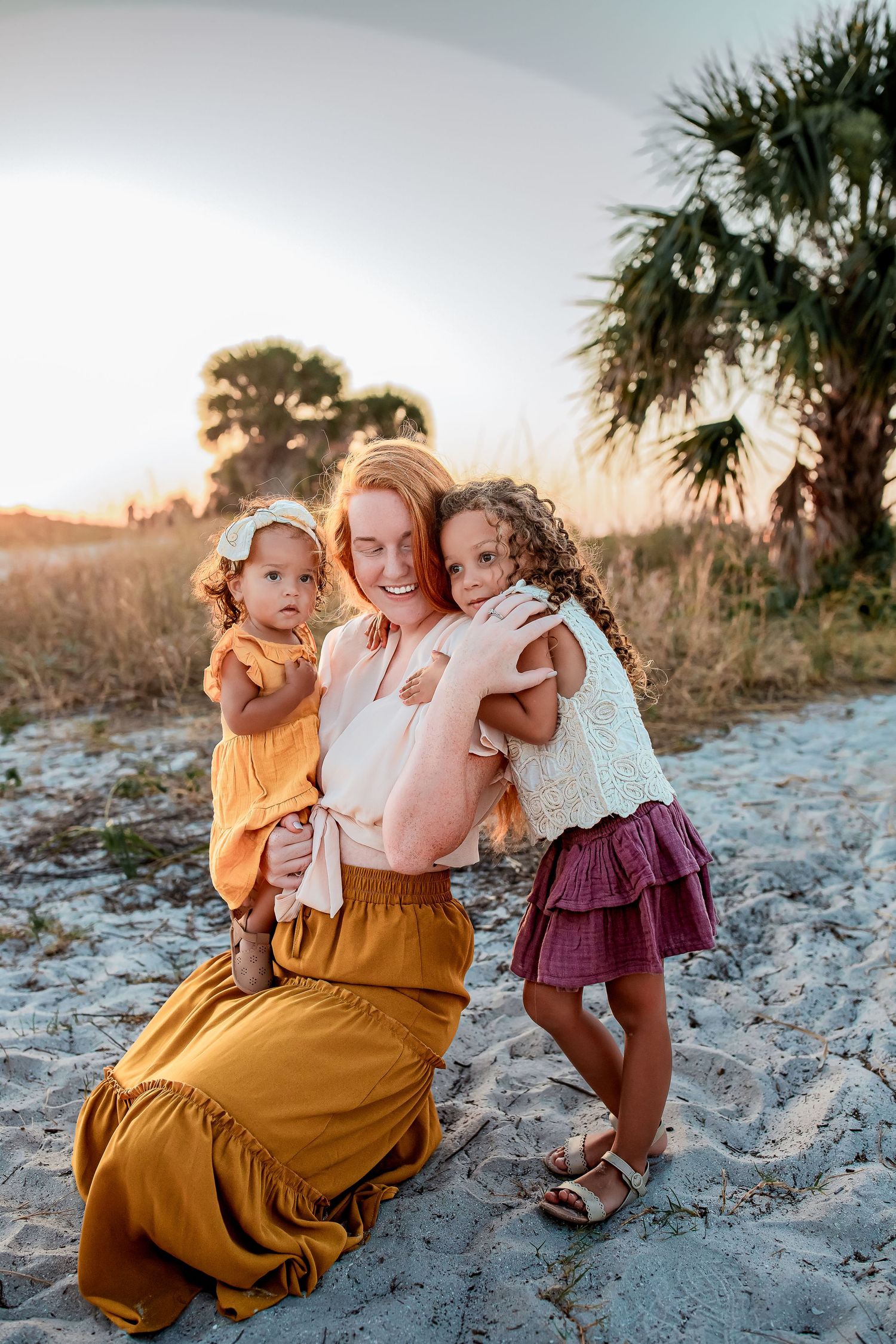 A redhead mother kneels on sandy beach holding two young daughters at golden hour sunset.