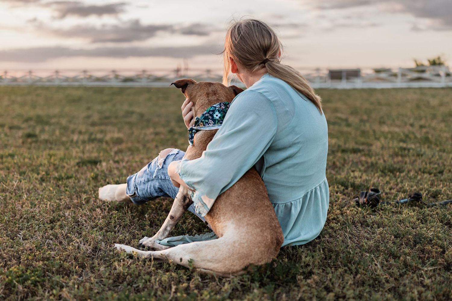 Woman sitting on grass with her dog at sunset, sharing a tender moment outdoors.