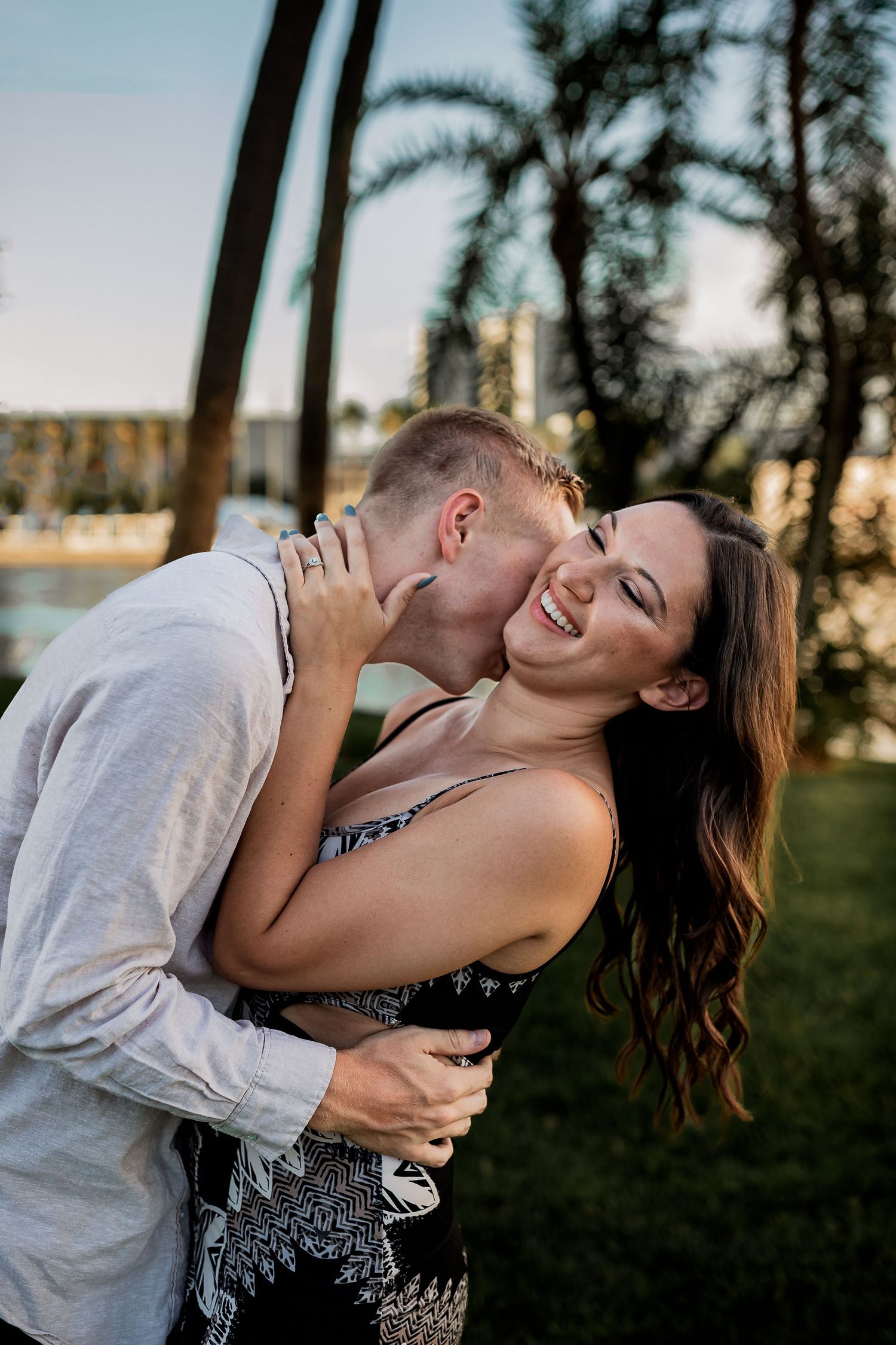 Man kisses woman on the cheek outdoors near palm trees in warm golden evening light.