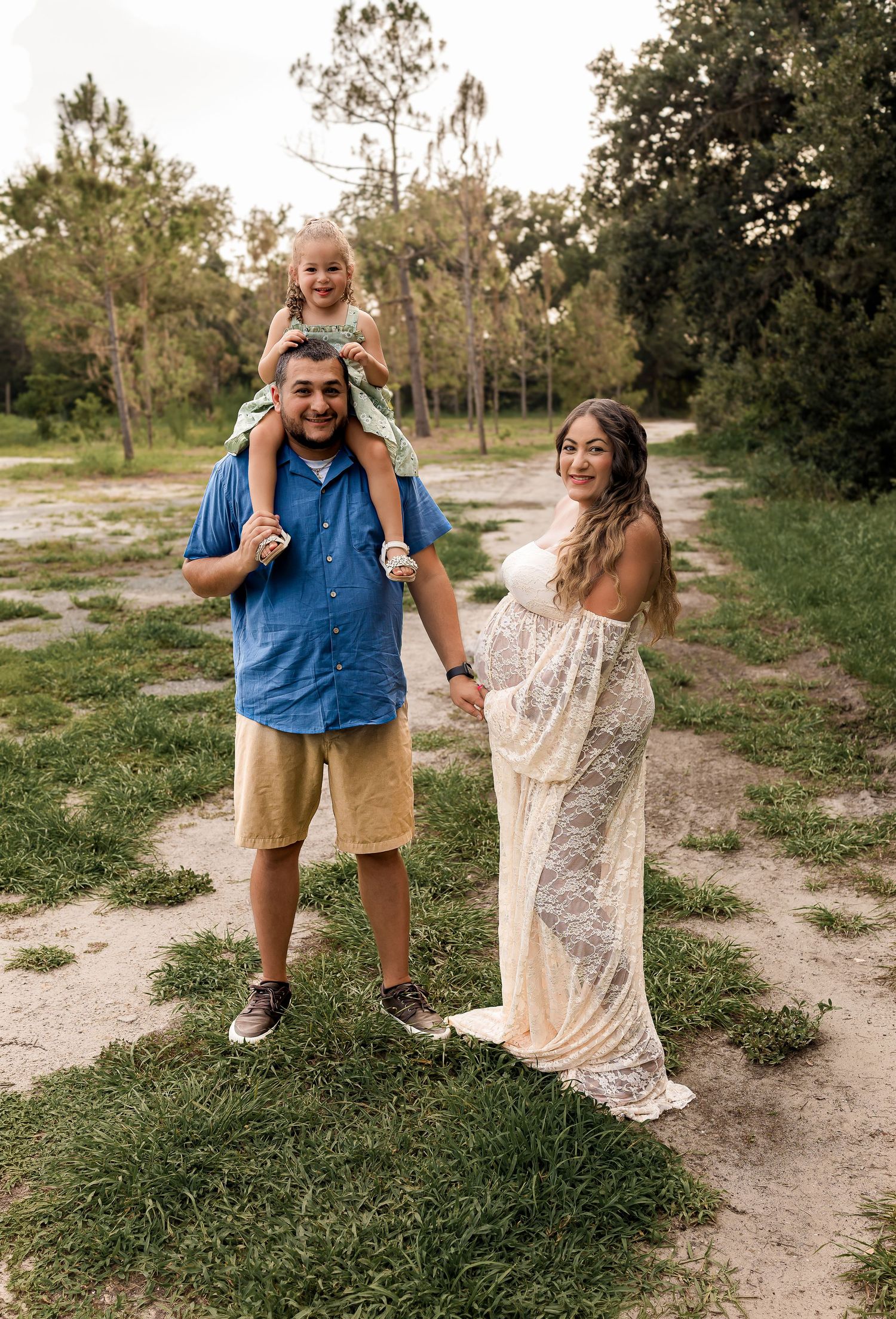 A family with a toddler on dad's shoulders and a pregnant mom in white lace dress outdoors.