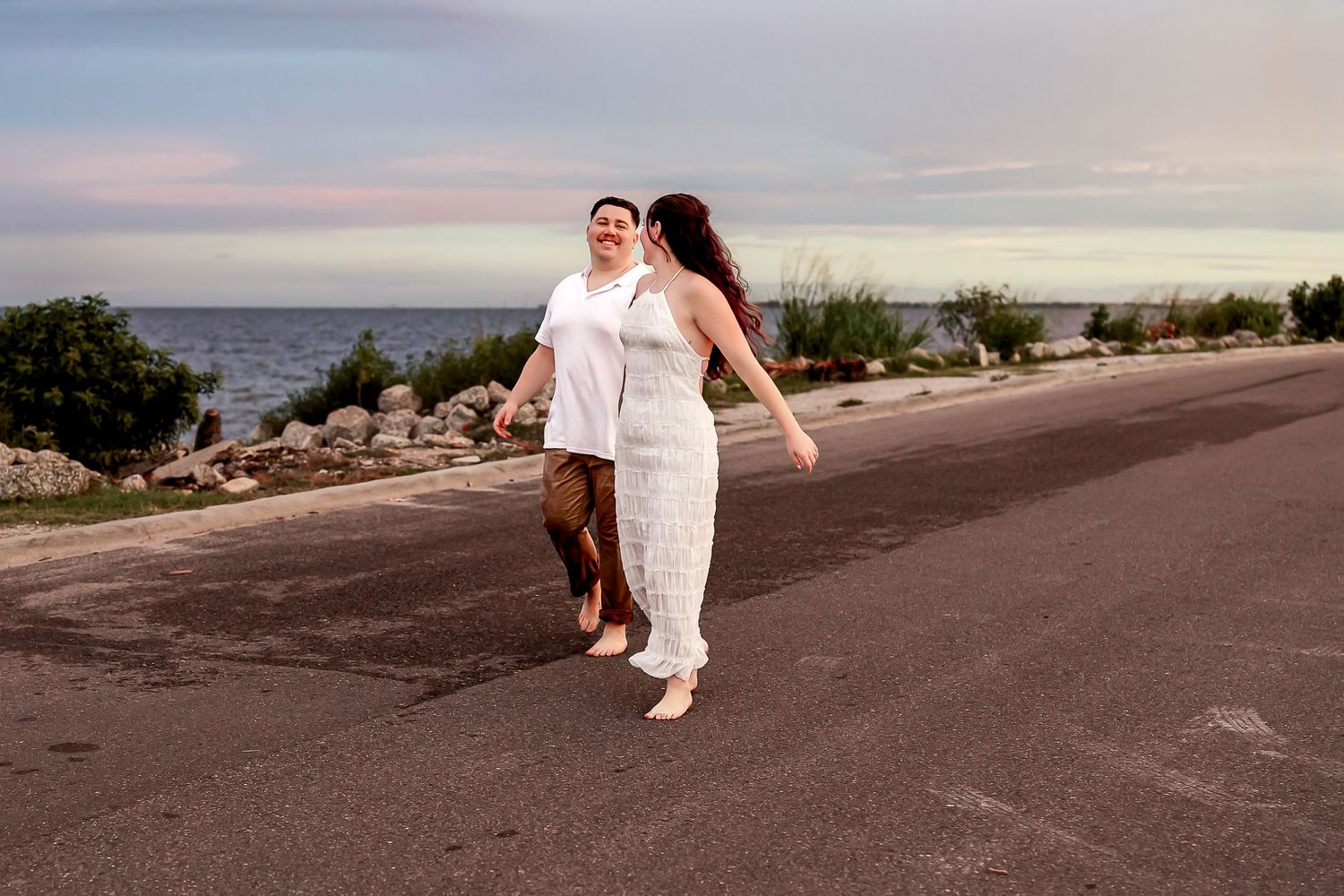 Couple dancing together on empty road near waterfront at dusk in casual white outfits.