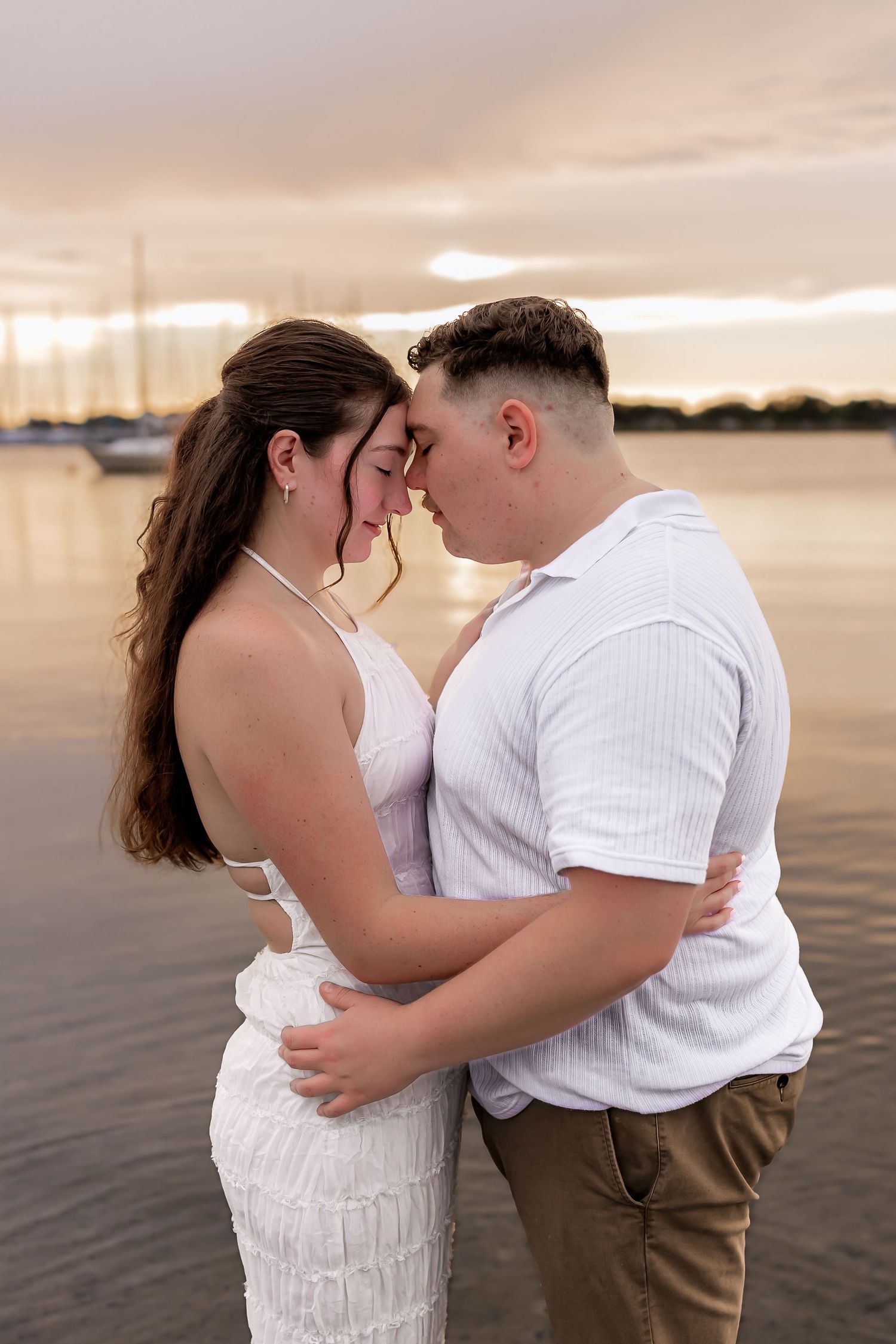 Romantic couple in white embraces forehead-to-forehead at waterfront during golden sunset.