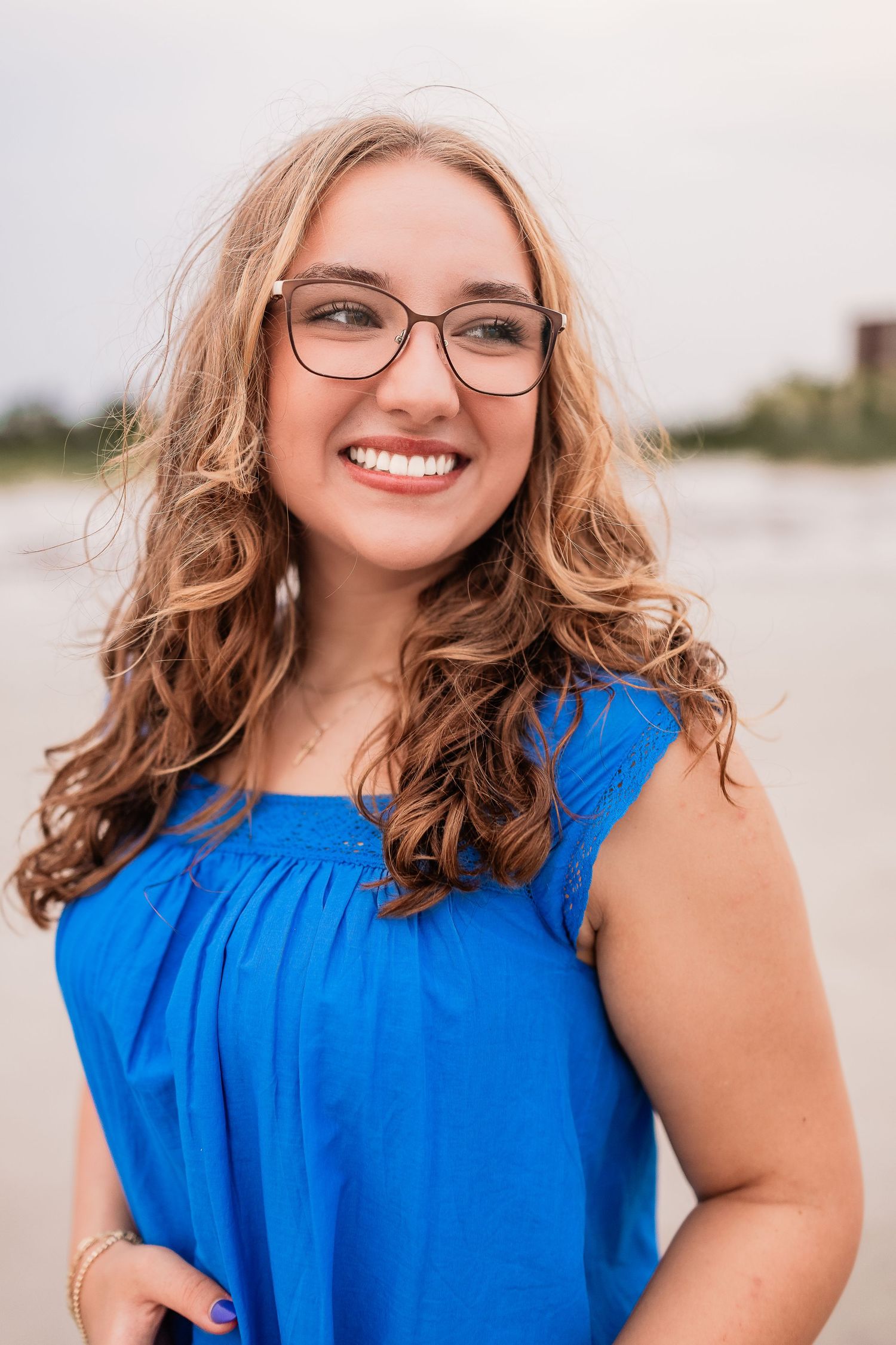 Cheerful teen girl with glasses and curly hair wearing blue top at beach.