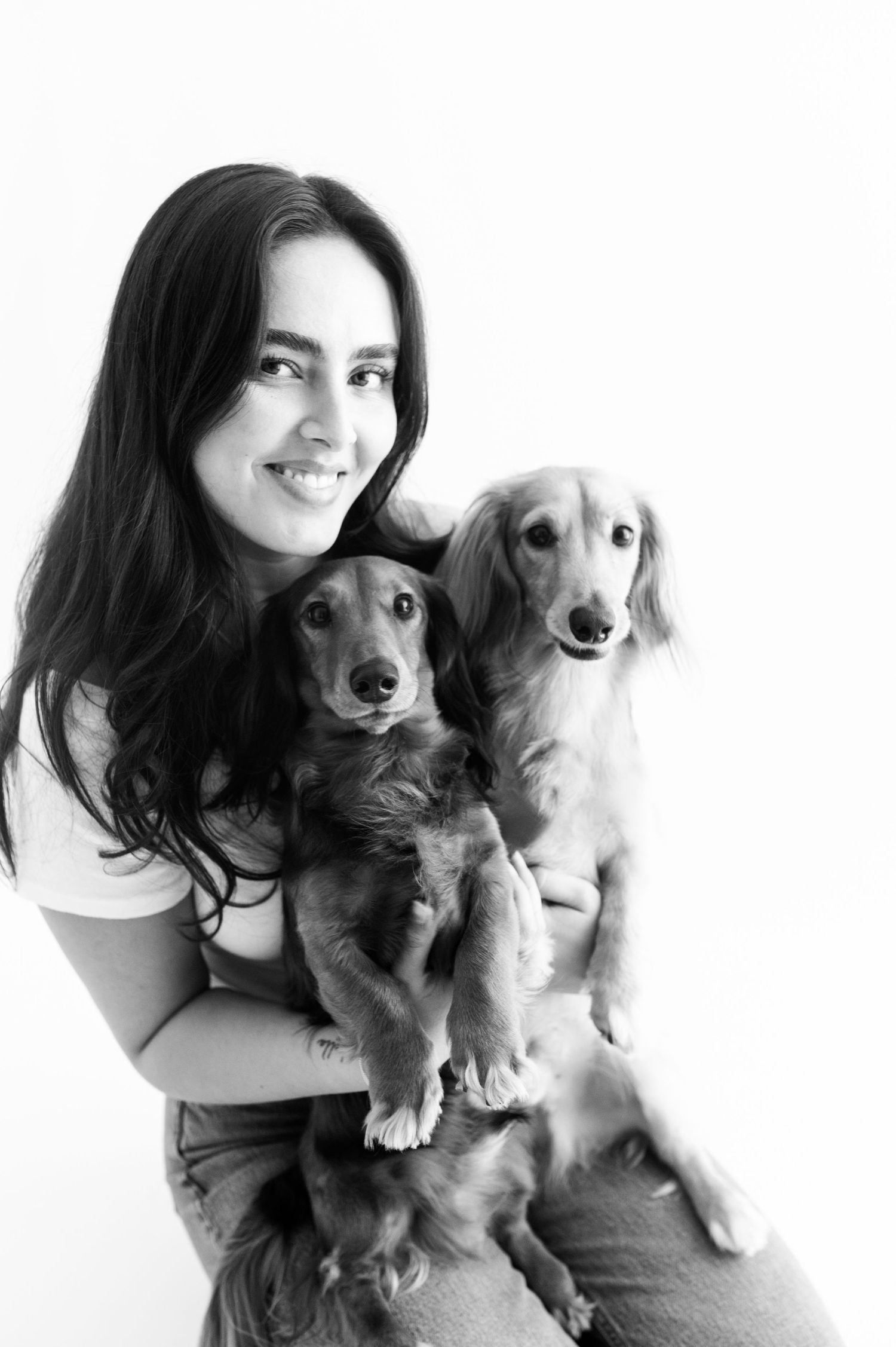Smiling woman holding two adorable long-haired Dachshunds in black and white portrait.