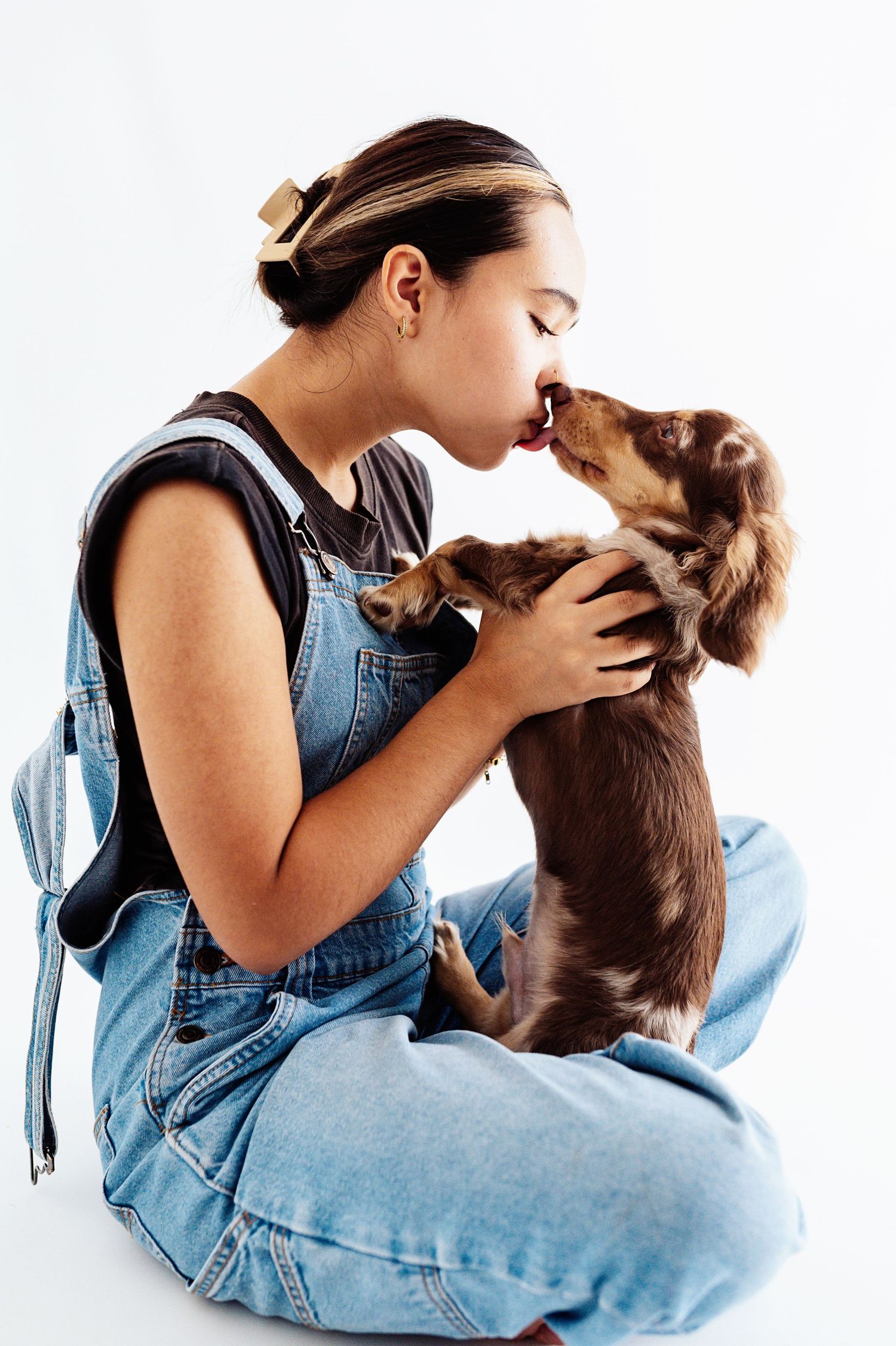A young girl in denim overalls being licked by a small dachshund puppy.