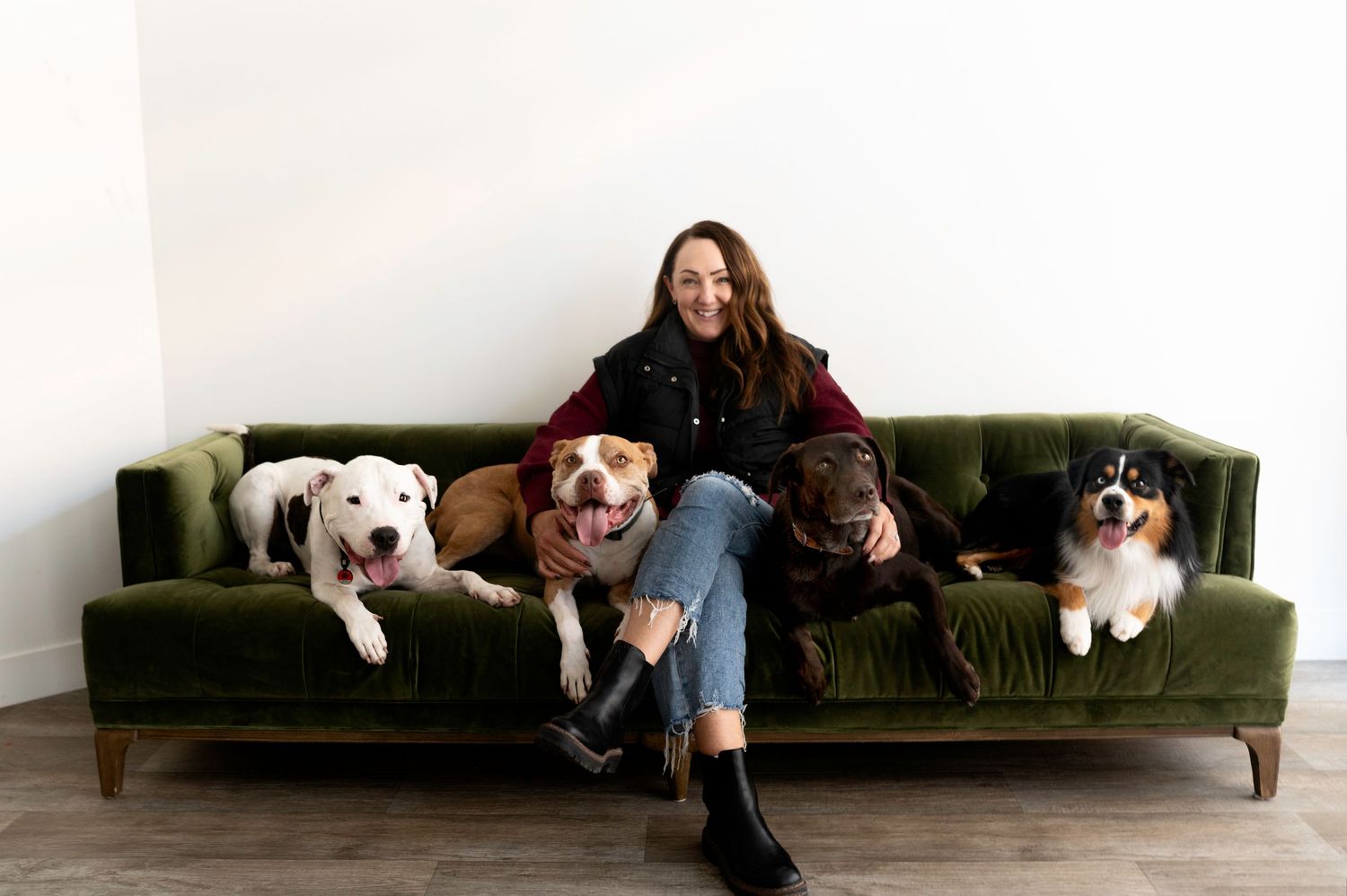 Smiling woman sitting on green velvet sofa surrounded by four large mixed breed dogs.