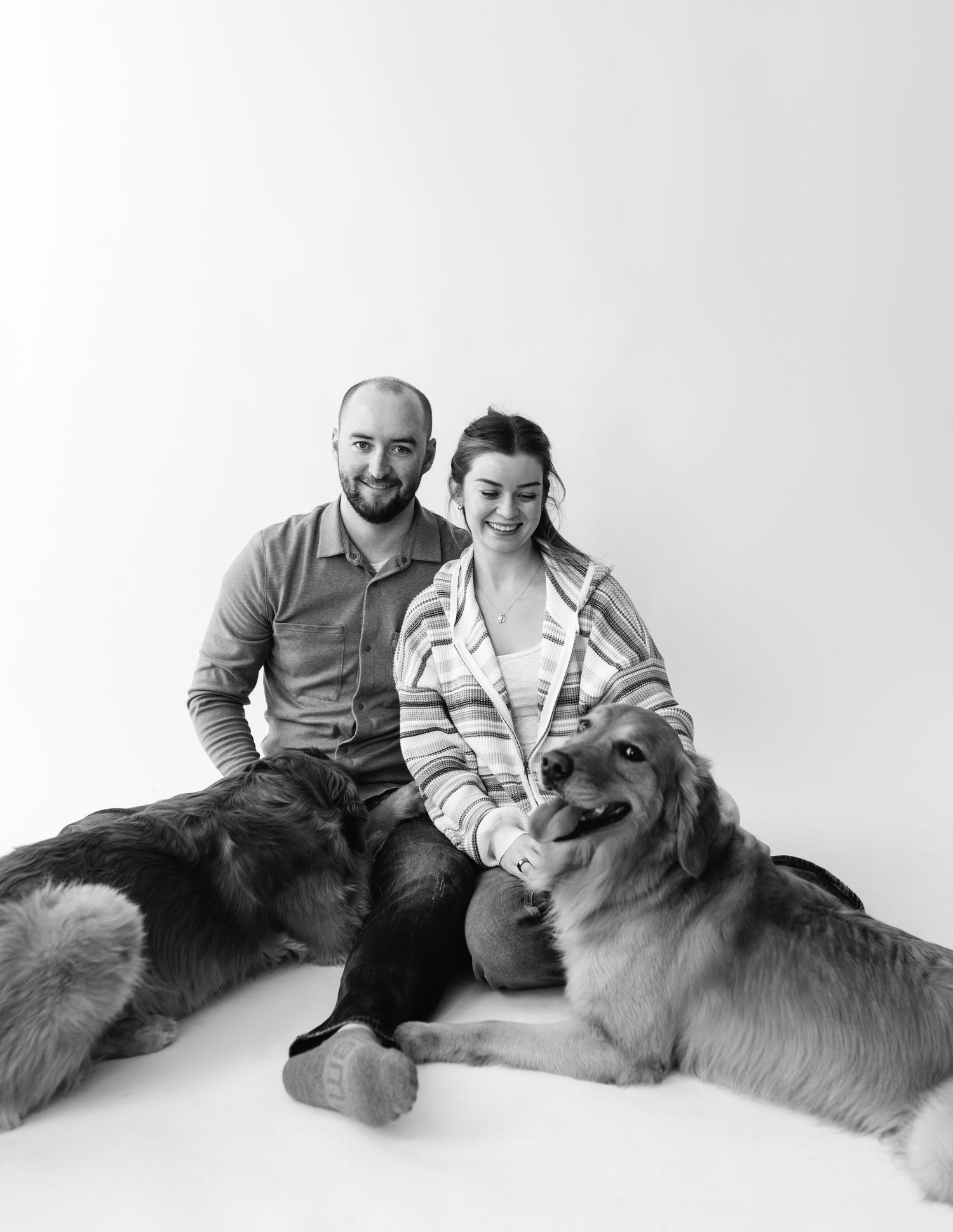 Black and white photo of smiling couple sitting with two large dogs on floor.