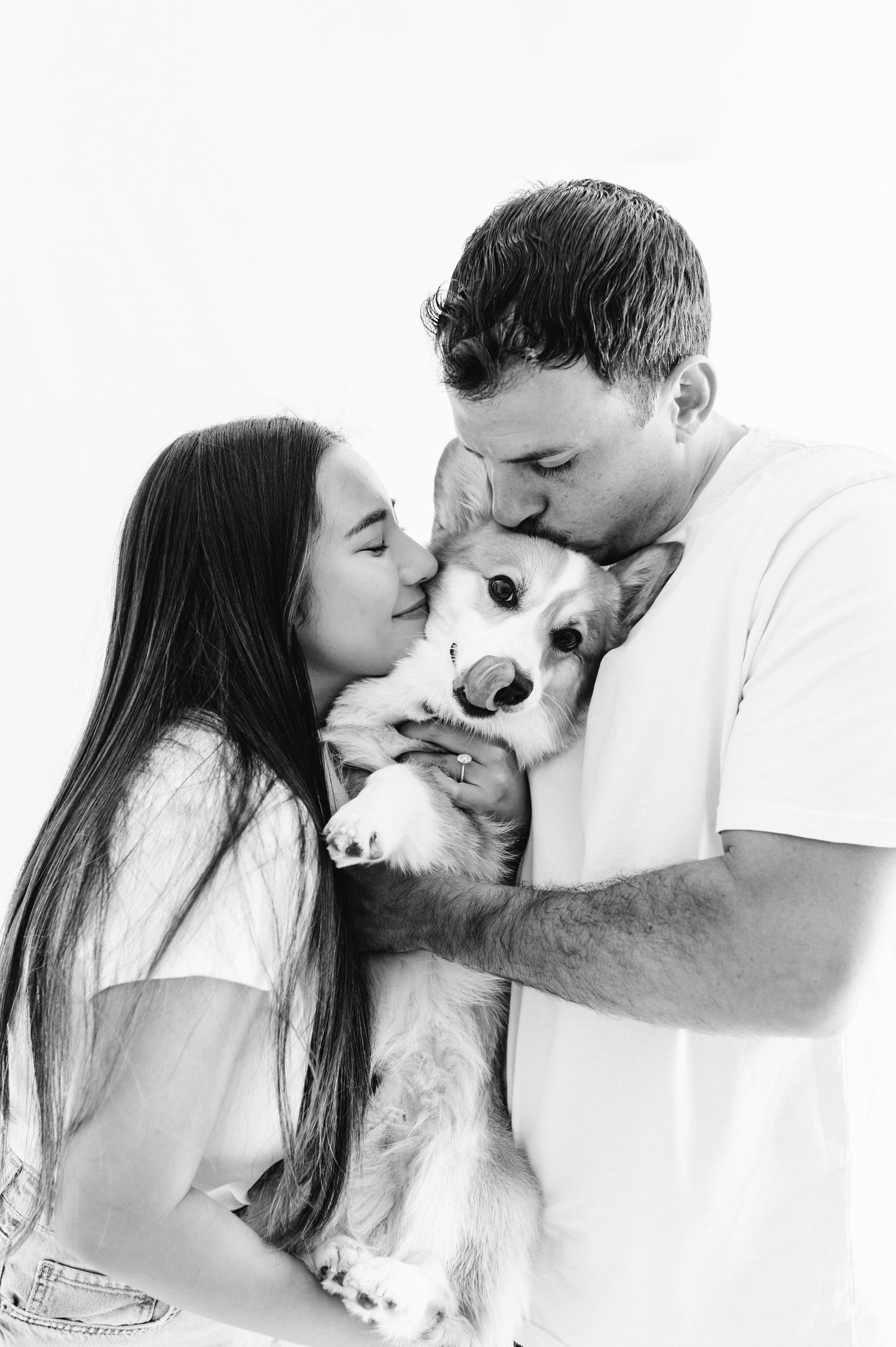 Black and white photo of couple kissing their Corgi dog on white background.
