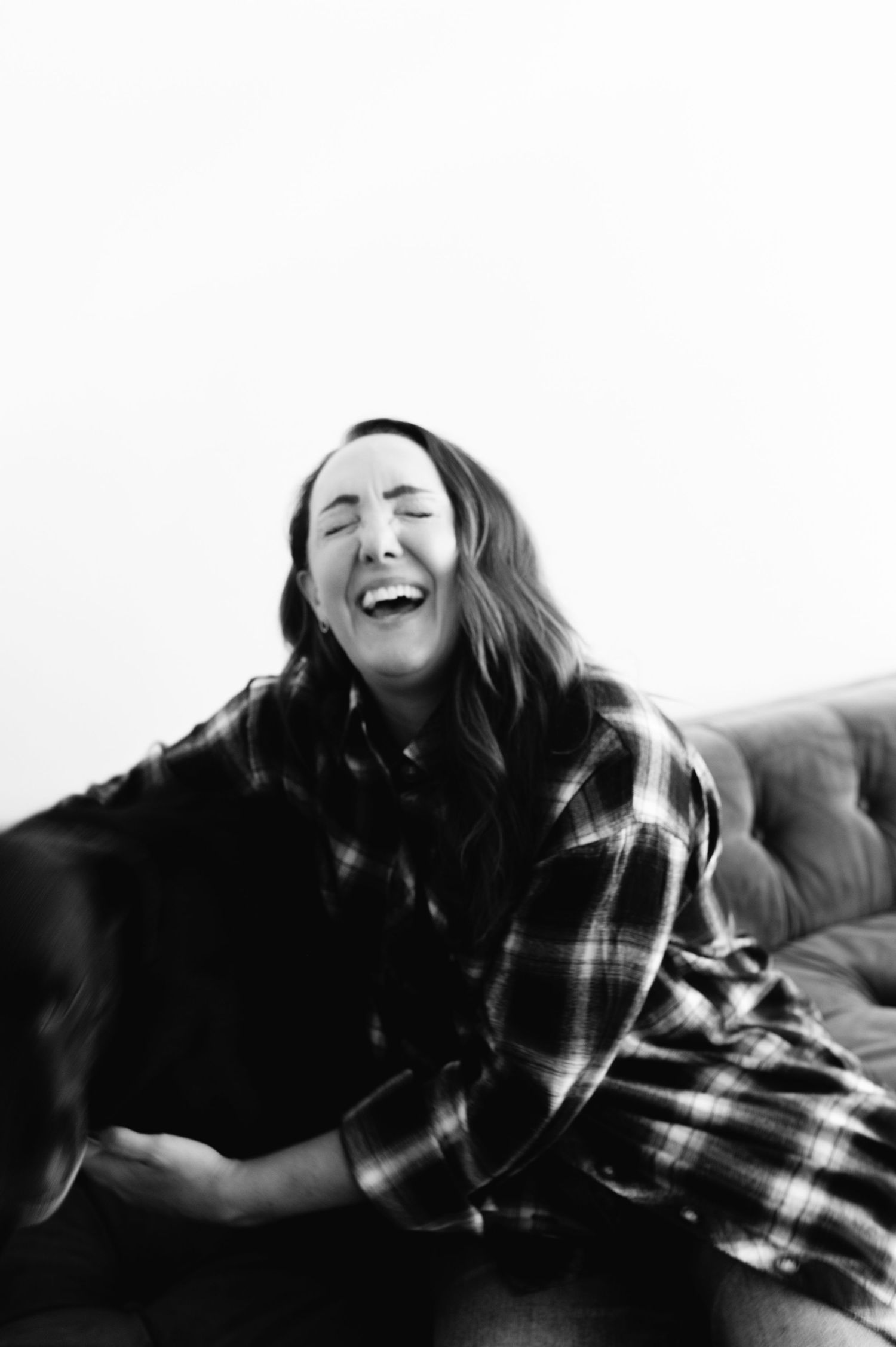 A woman laughing joyfully while sitting on a couch in a plaid shirt, black and white photo.