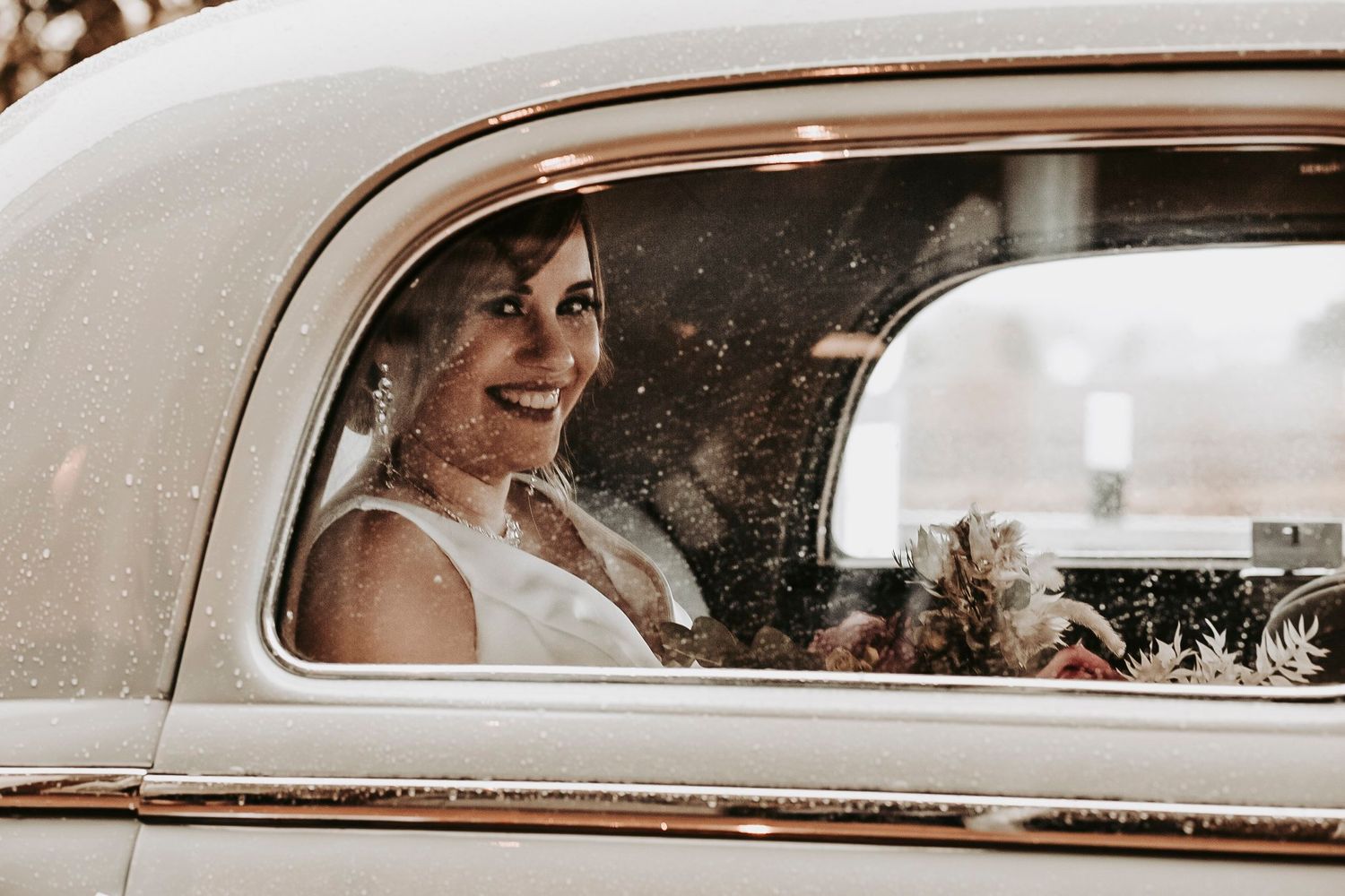 Smiling bride in a vintage white car, wearing a sleeveless wedding dress and veil.