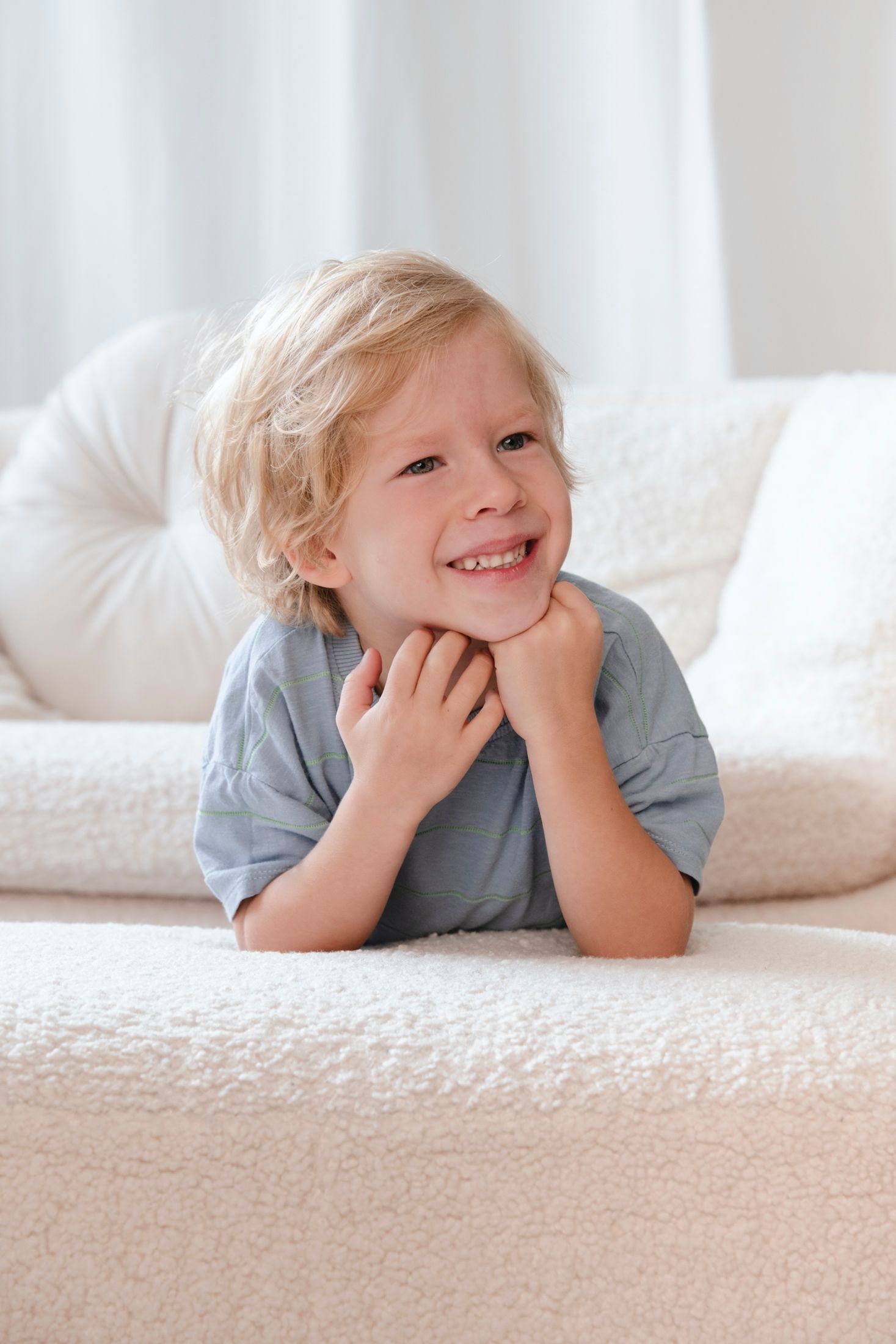Smiling blonde boy lying on plush cream sofa with hands under chin in bright room.