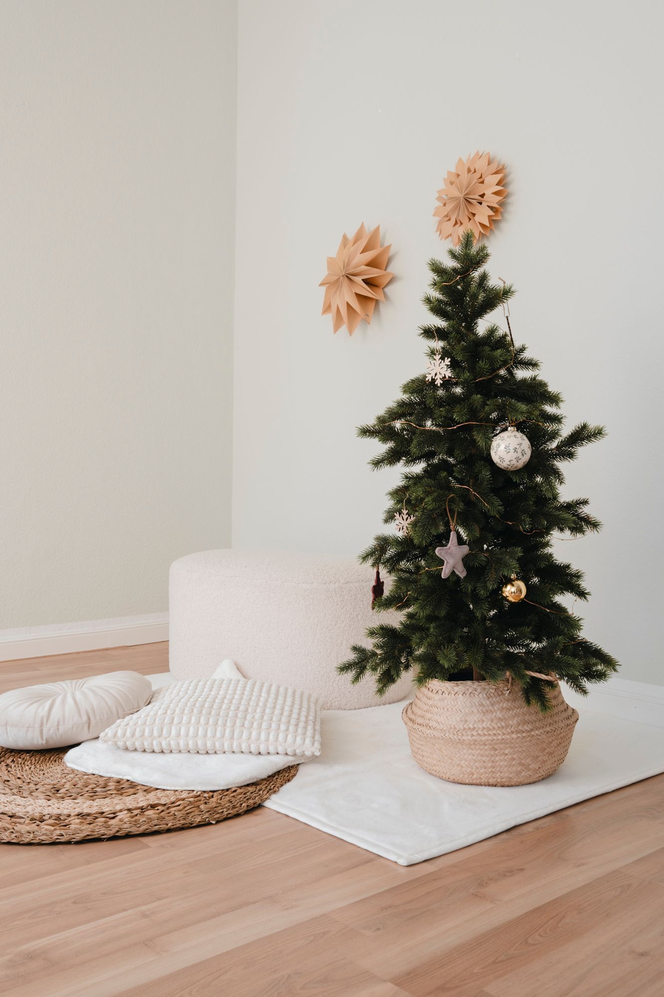 Minimalist Christmas tree in wicker basket with gold ornaments, beside white cushions on rattan tray, paper star wall decor.