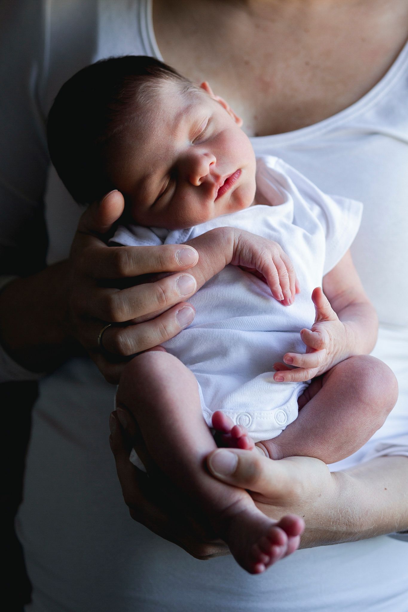 Newborn baby sleeping peacefully in parent's arms wearing a white onesie.