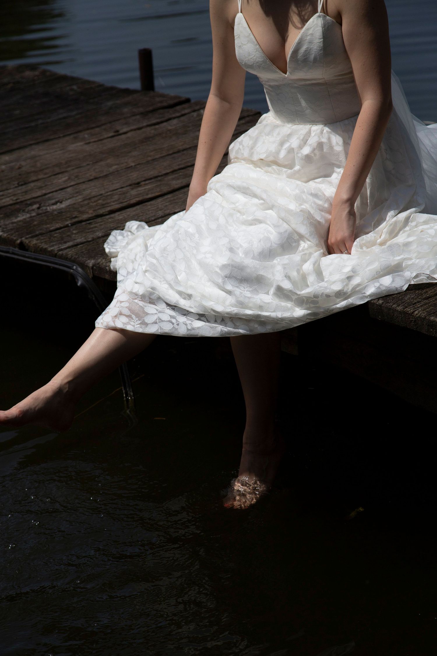 Bride in white dress sits barefoot on a wooden dock with feet dangling over the water in a serene outdoor setting.