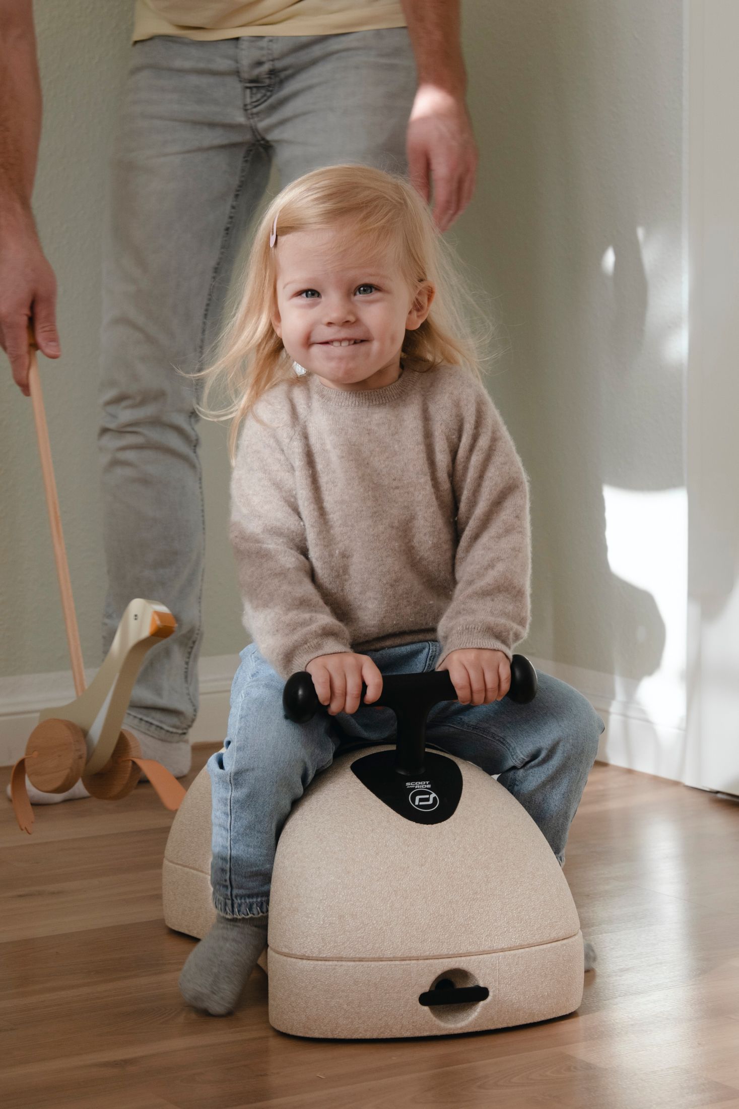 Happy blonde toddler girl riding sit-on toy with adult standing nearby on wooden floor.
