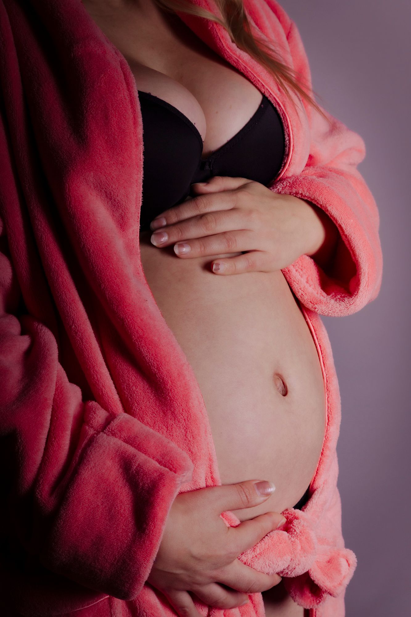 Close-up of pregnant woman in pink robe and black bra gently cradling her bare belly.