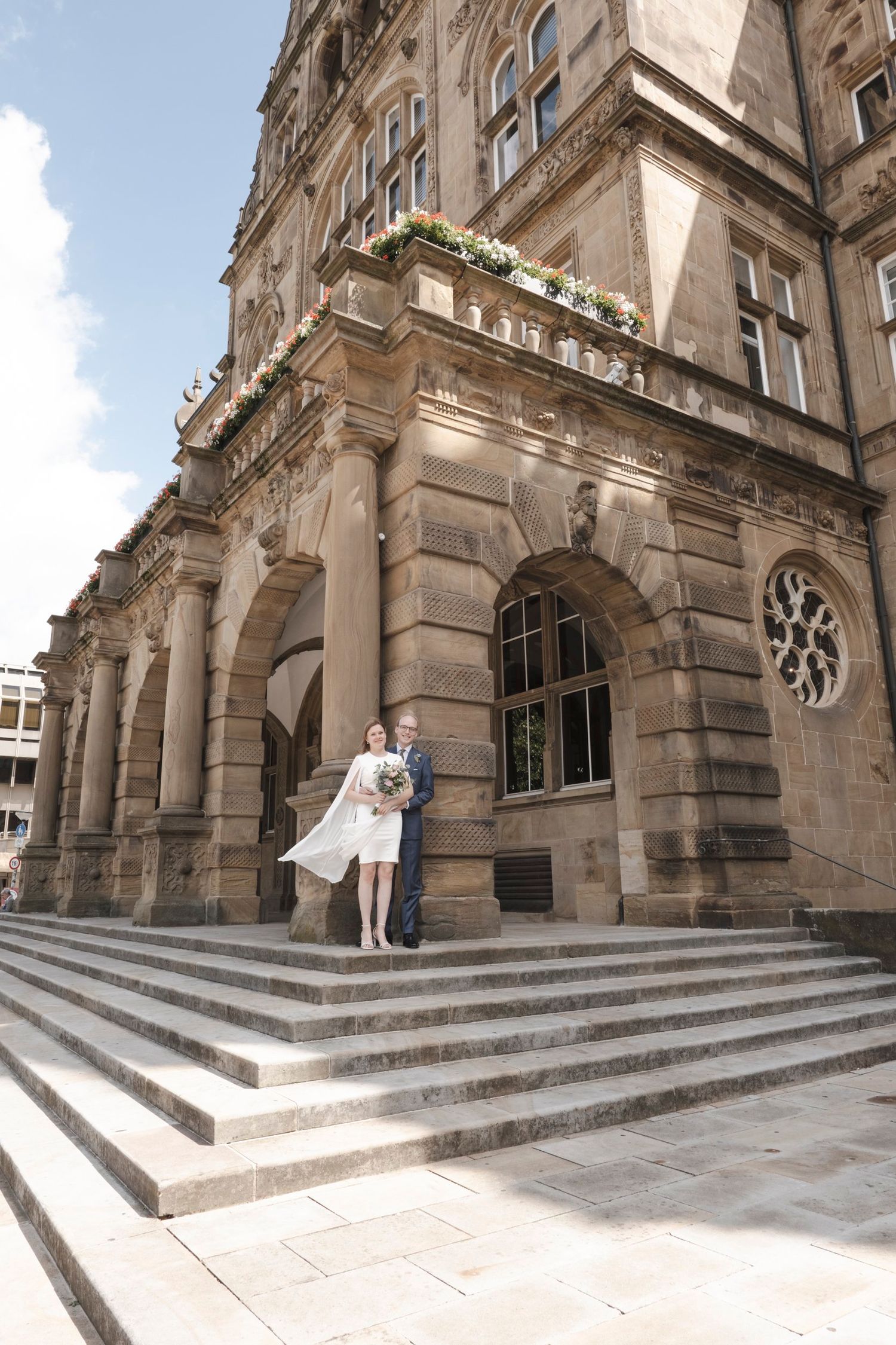 Elegant wedding couple posing on steps of ornate historic stone building under blue sky.