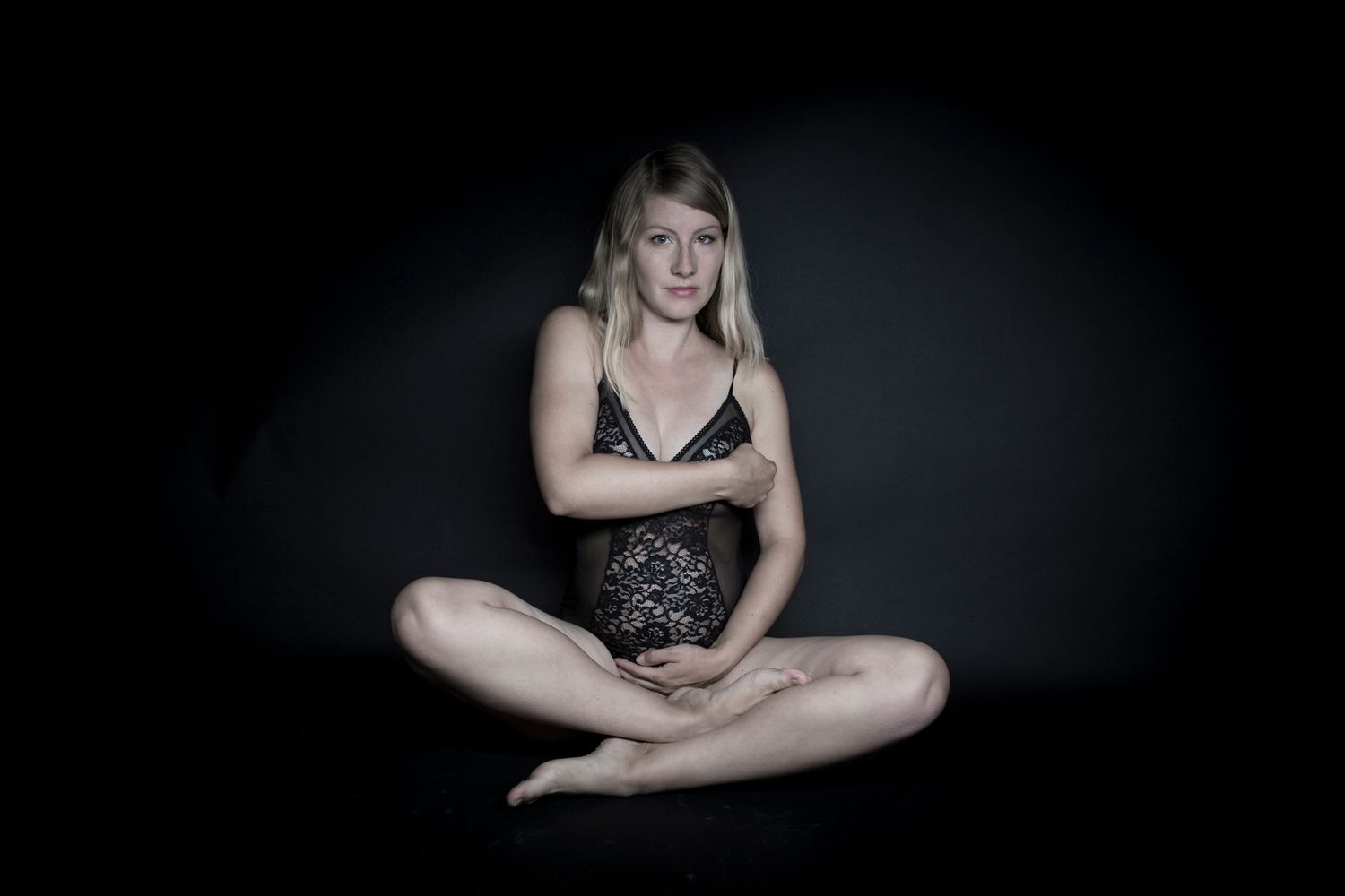 Pregnant blonde woman sitting cross-legged in black lace lingerie against a dark background.