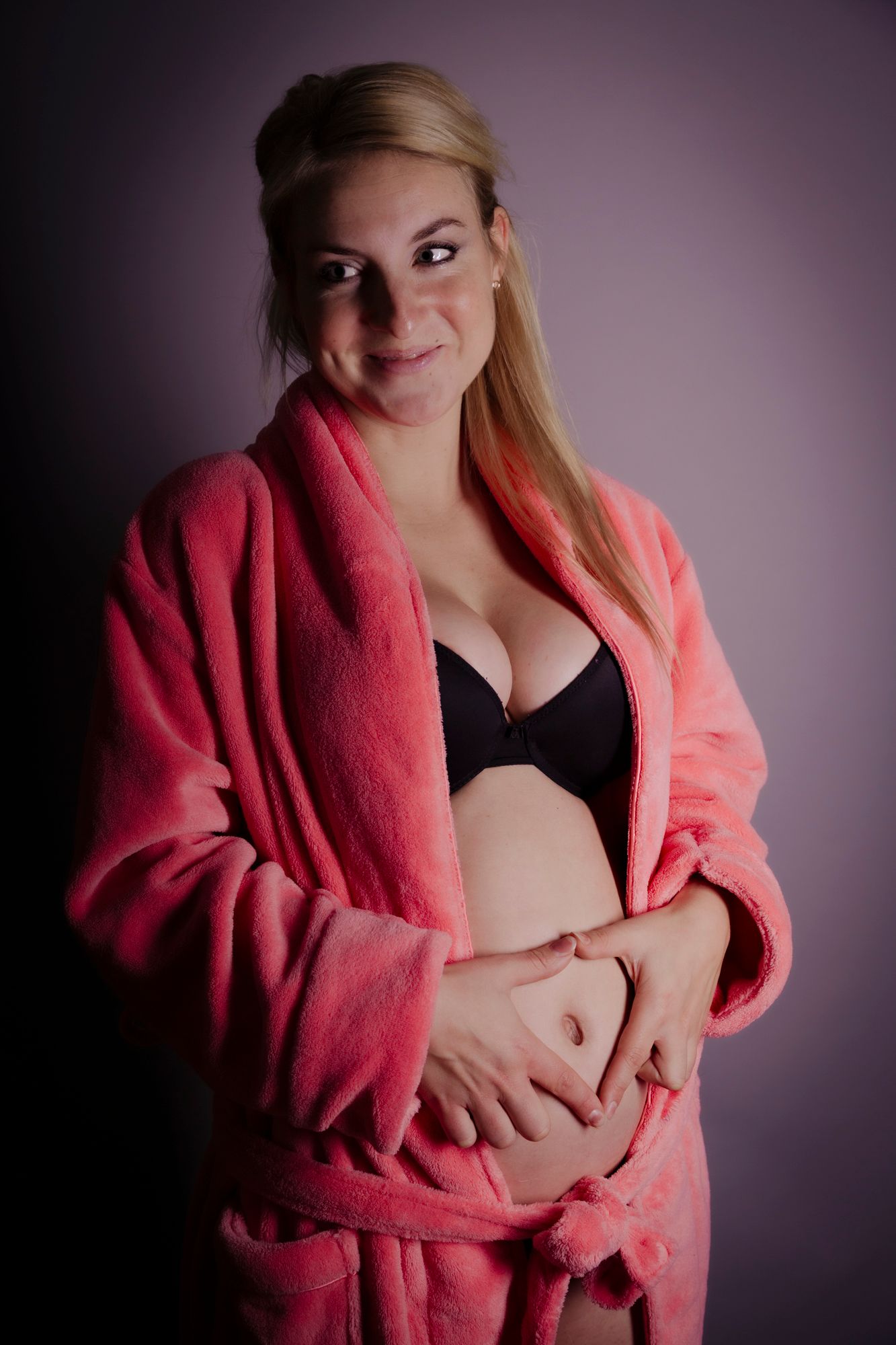 Pregnant blonde woman in open pink robe smiling while holding her bare rounded belly.