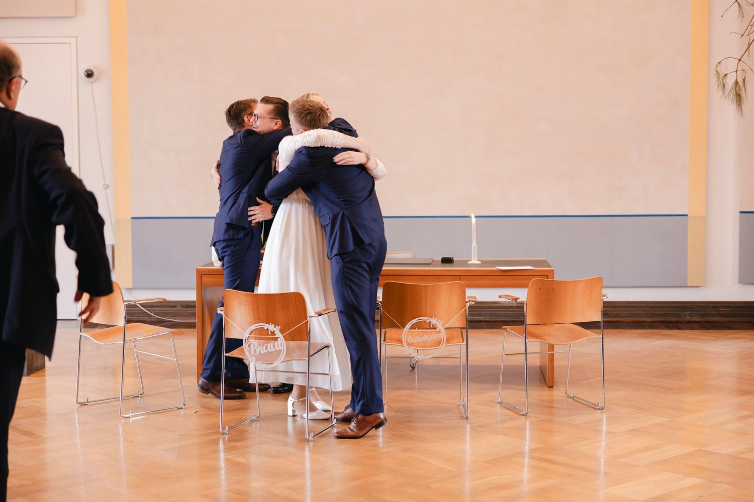 Bride and groom share a warm embrace with a guest after wedding ceremony indoors.