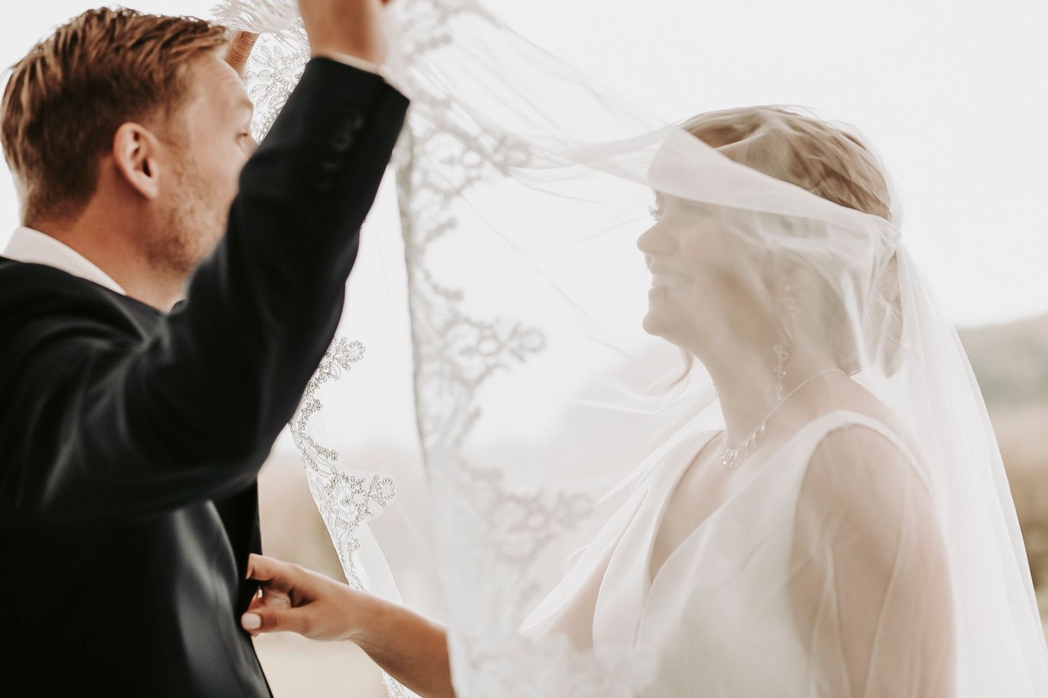 Groom lifting bride's lace veil in a soft, romantic wedding portrait with bright backlighting.