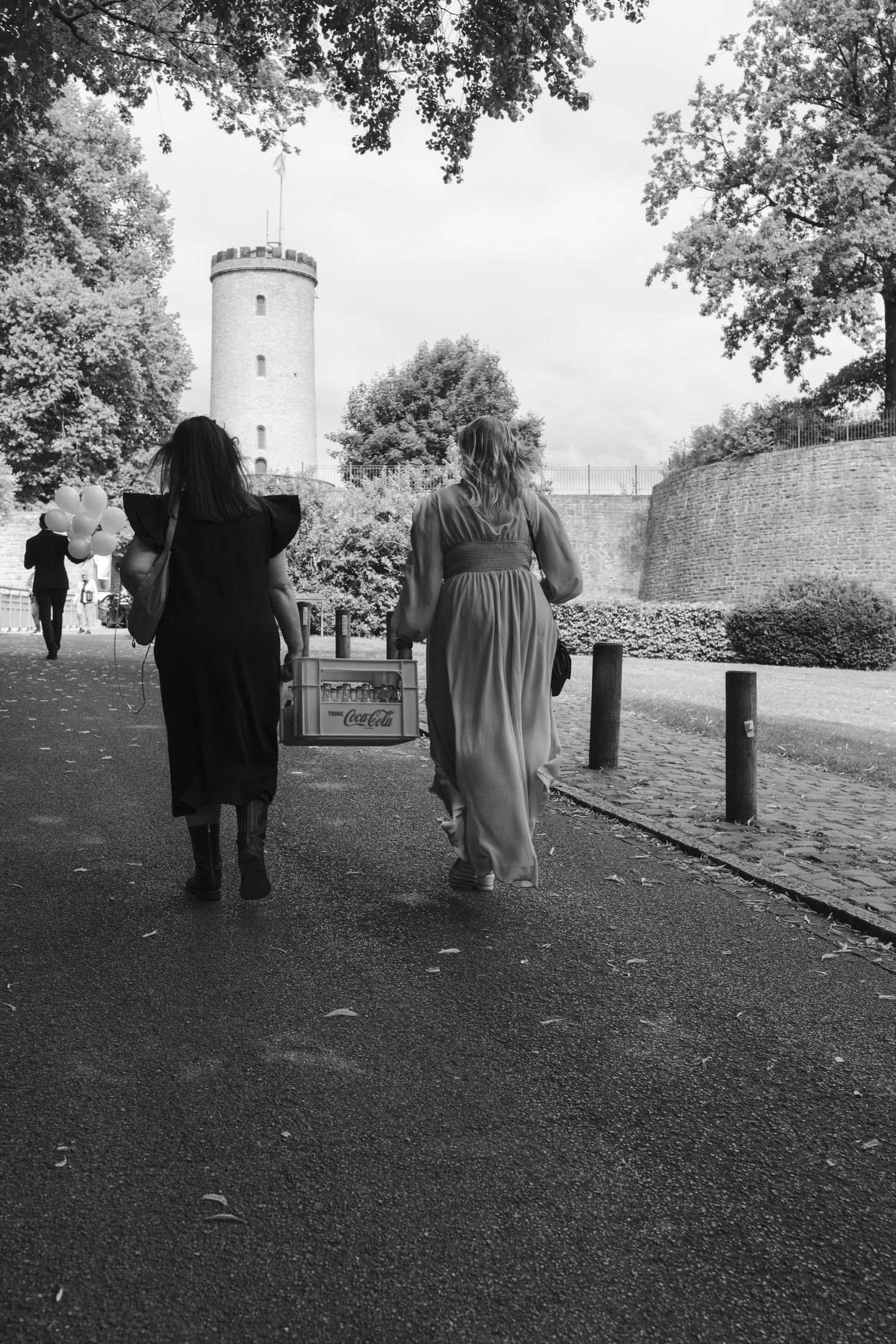 Black and white photo of two women in formal dresses walking toward a historic white tower amid trees.