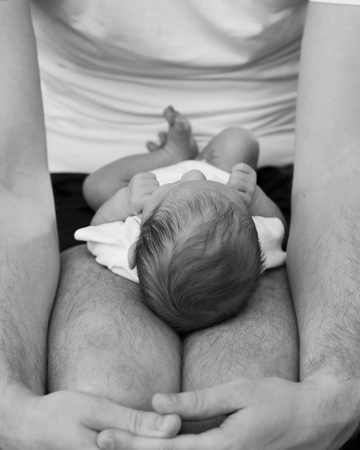 Black and white photo of newborn baby resting on father's lap in diaper.