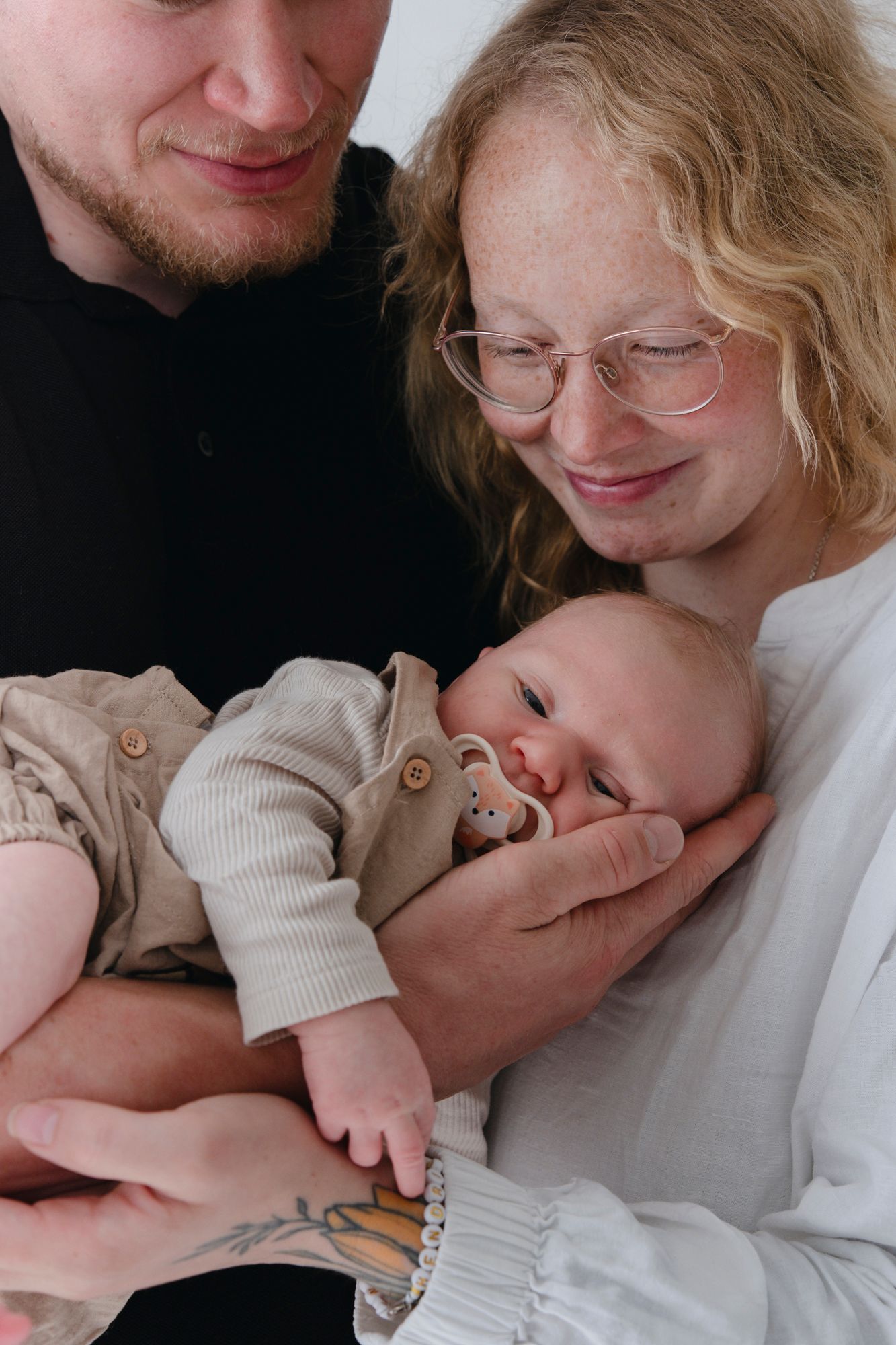 Happy young parents smiling lovingly at their newborn baby with pacifier held between them.
