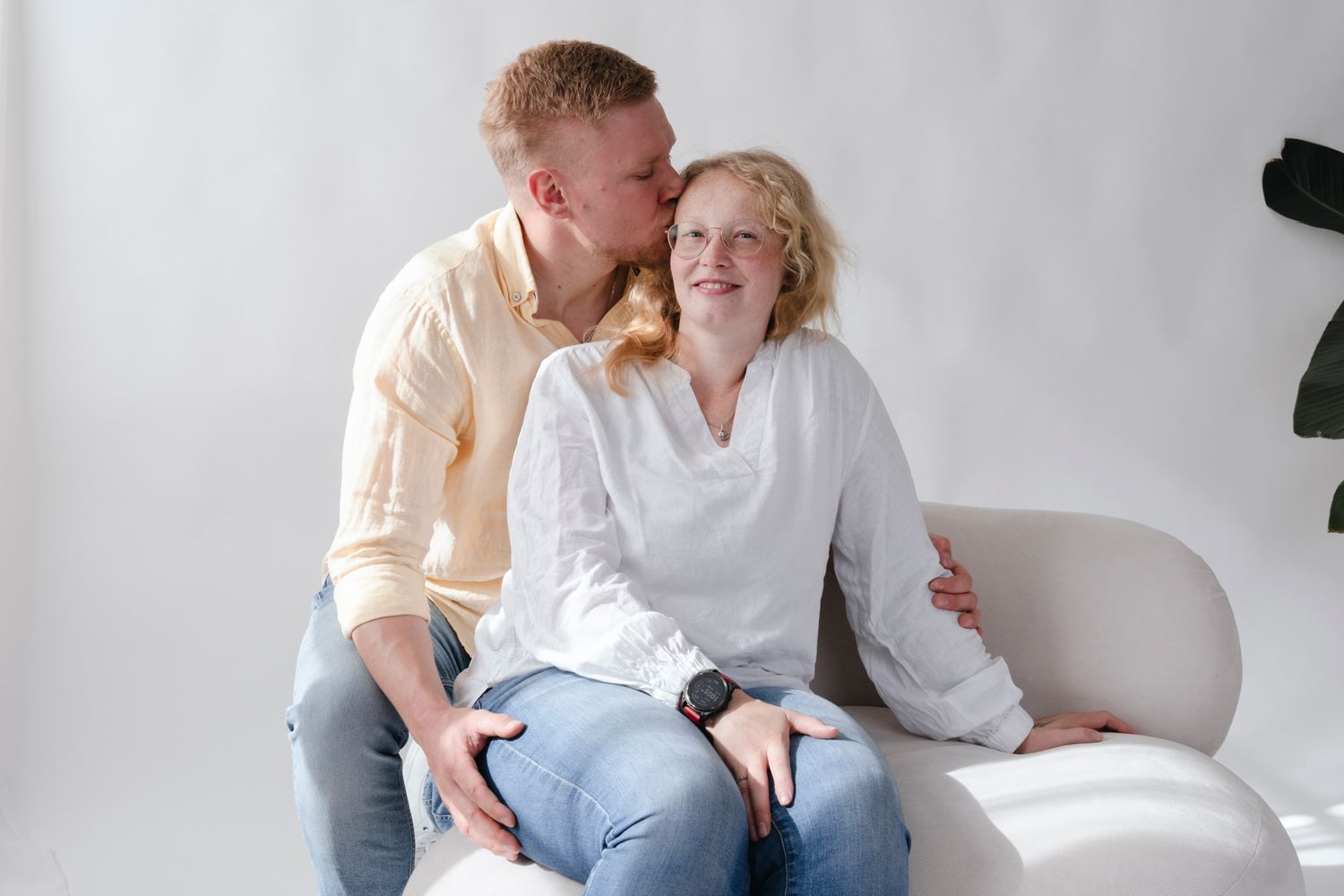 Happy couple sitting on a white sofa, man kissing smiling woman on the cheek in bright room.