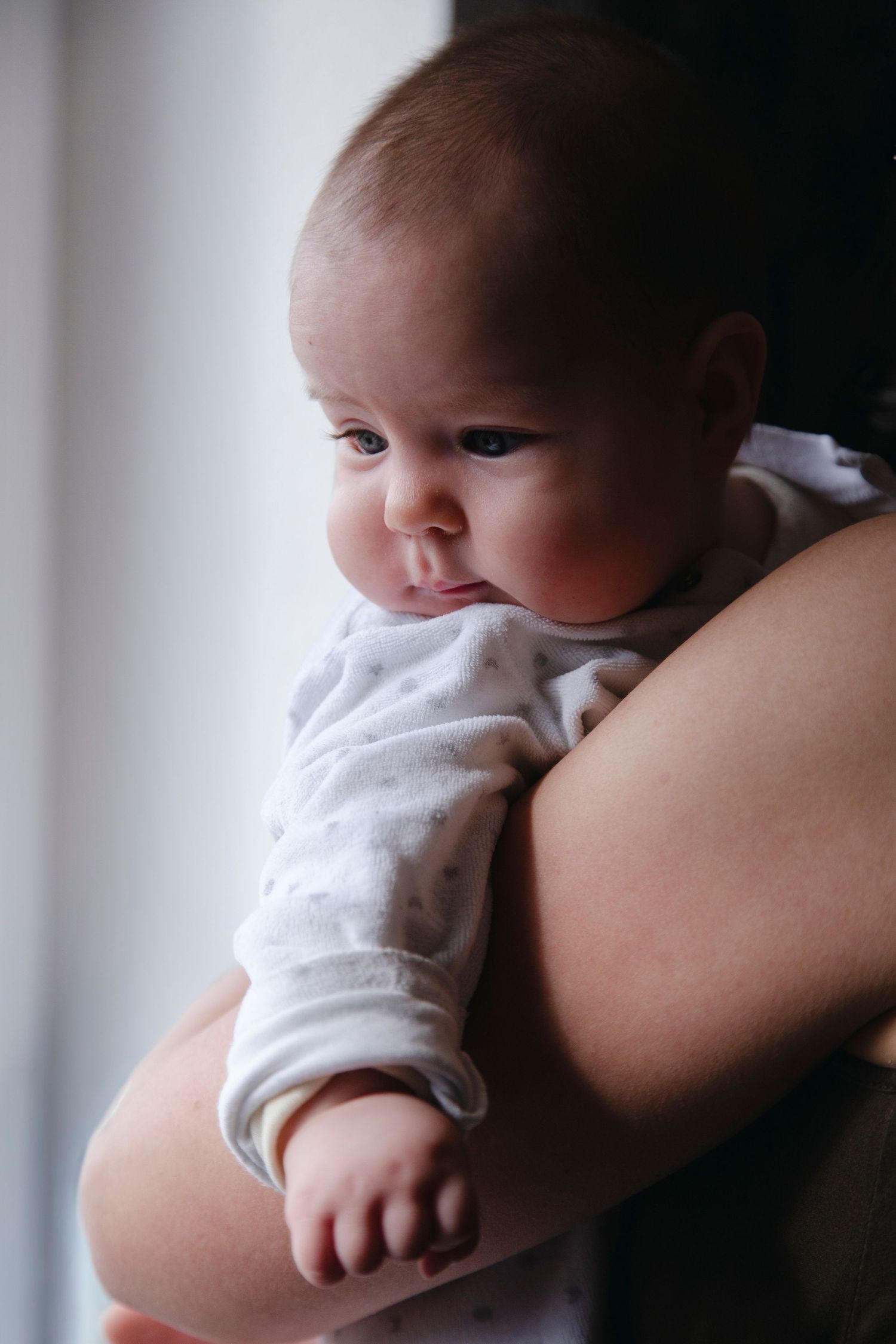 Alert baby in white outfit resting on parent's shoulder near a bright window.