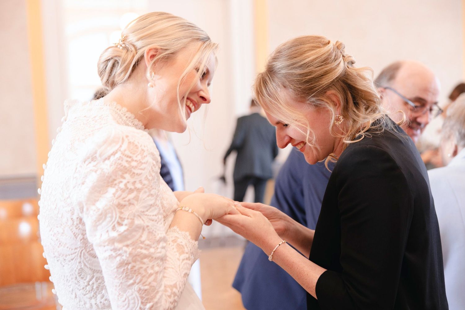 Bride in lace dress laughing joyfully with female guest admiring wedding ring after ceremony.